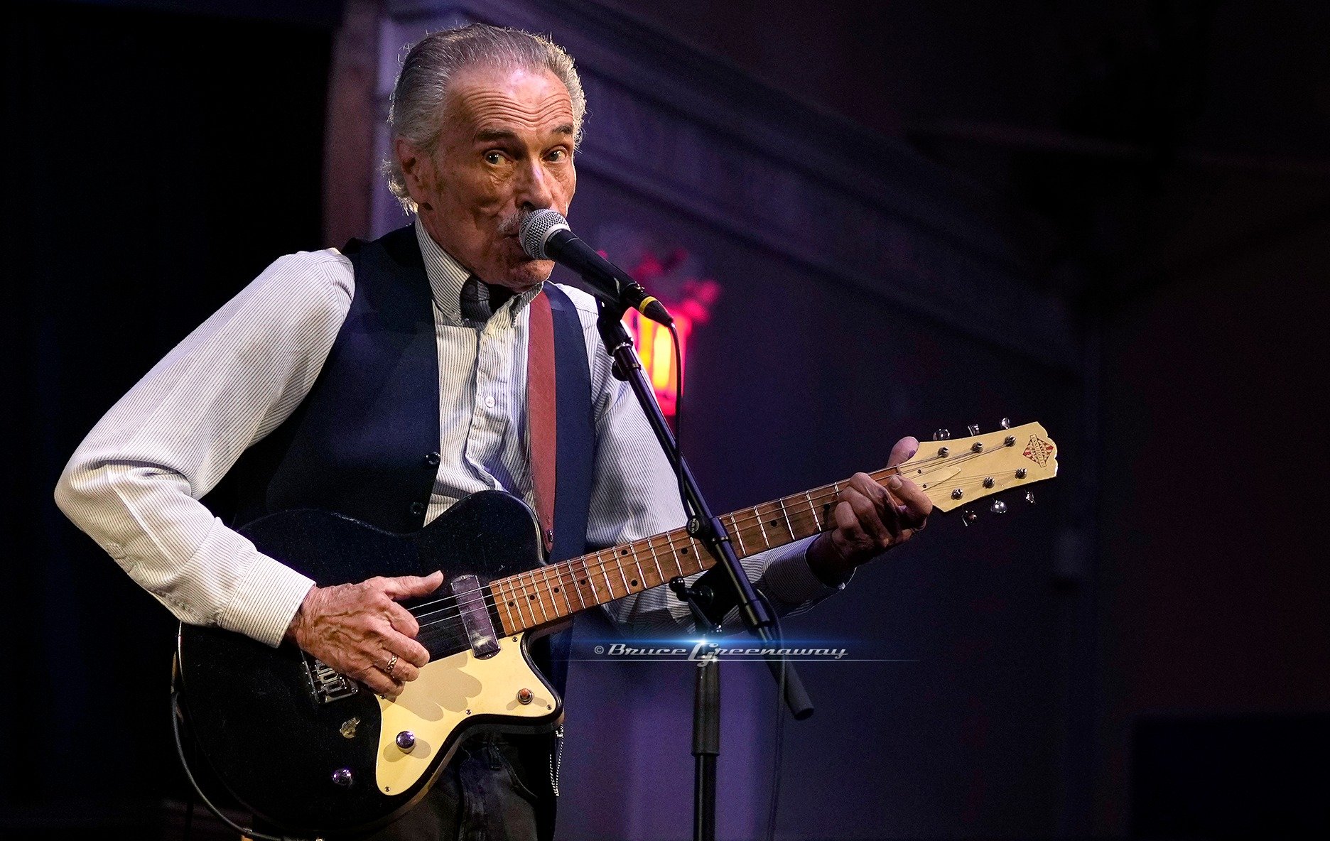 An elderly man with gray hair singing into a microphone while playing a black electric guitar on stage with dark background.