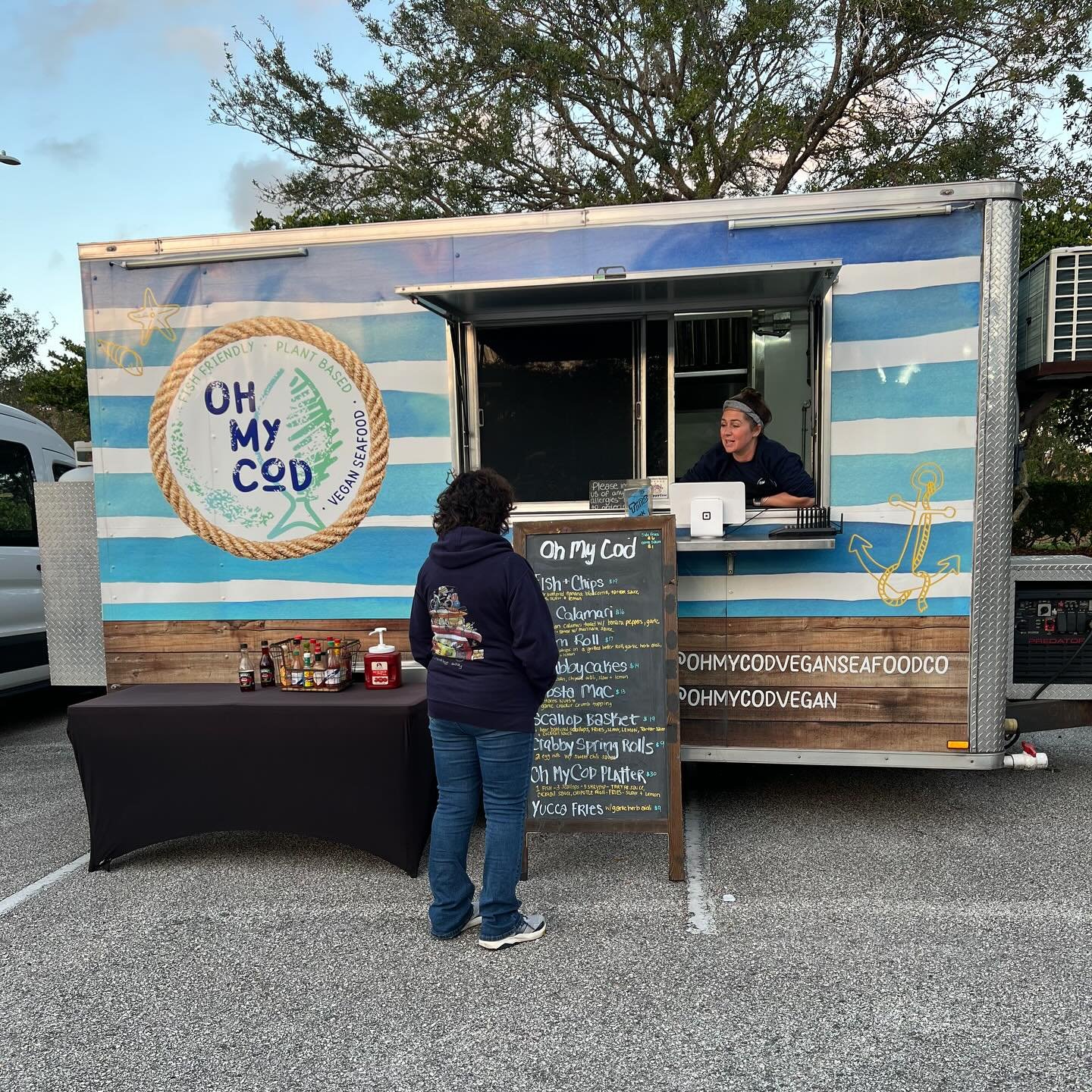 A food truck named 'Oh My Cod' is serving vegan fish and seafood dishes. A woman is ordering at the counter, while the staff inside behind the window is preparing the order. The truck is decorated with maritime-themed graphics, including an anchor and a rope circle logo.