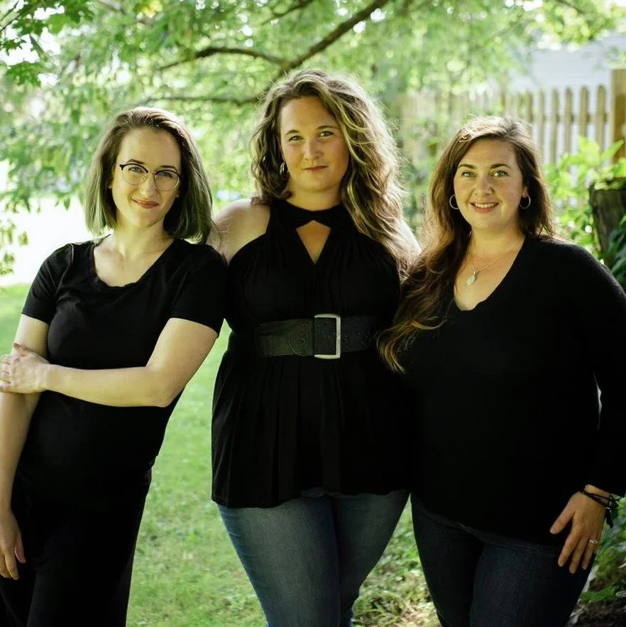 Three women standing outdoors in a green garden, smiling at the camera. They are dressed in black tops and jeans, with trees and a wooden fence in the background.