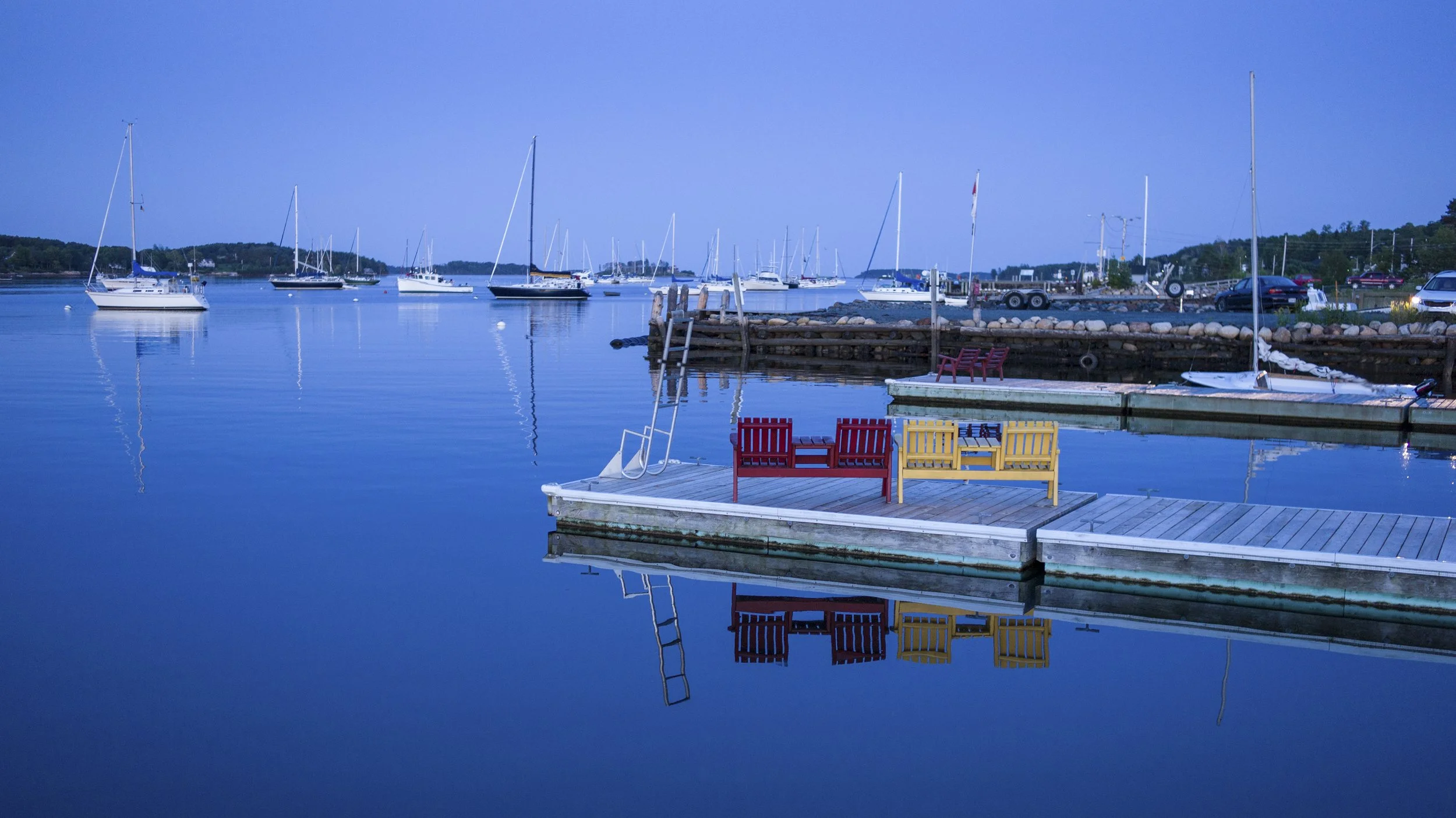 A serene marina scene at dusk with sailboats floating on calm blue water, colorful benches on a dock, and a clear evening sky.