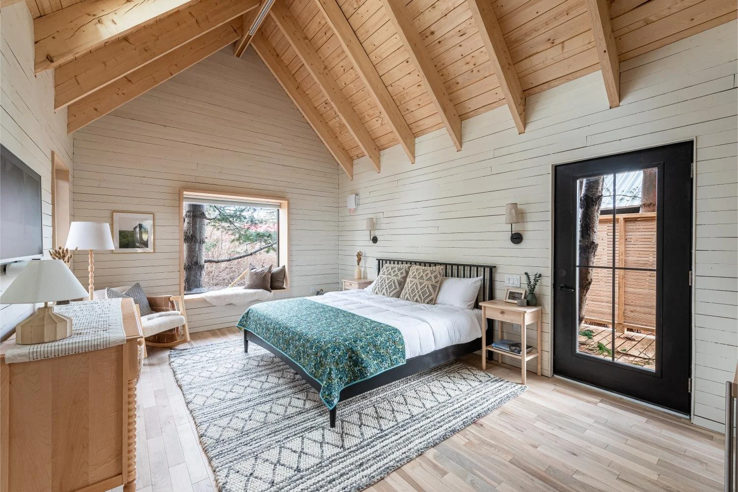 A bedroom with a vaulted wooden ceiling, white shiplap walls, a large bed with decorative pillows and a blue quilt, two bedside tables with lamps, a small white sofa by the window with cushions, and a glass door leading outside. There's a window with a view of trees and a rug on a wooden floor.