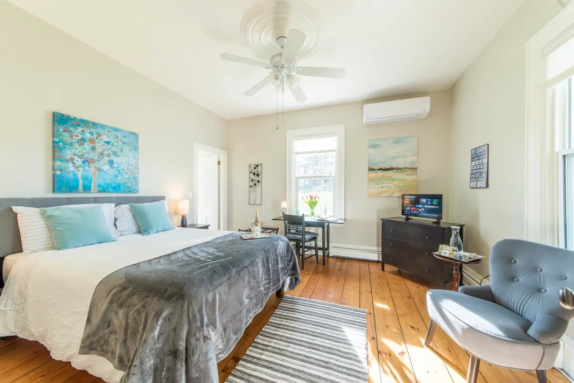 Bright bedroom with a large bed, painting above, small table with glases and bottles, small TV on dresser, desk with flowers, cozy chair, and large windows letting in sunlight.