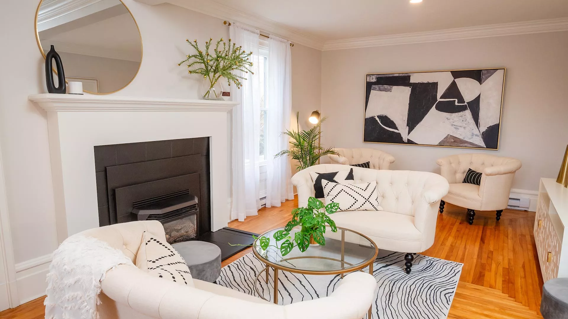 Living room with white sofas, black and white pillows, a glass coffee table with green plants, a zebra-print rug, abstract black and white artwork, a fireplace with black trim, and large windows with white curtains.