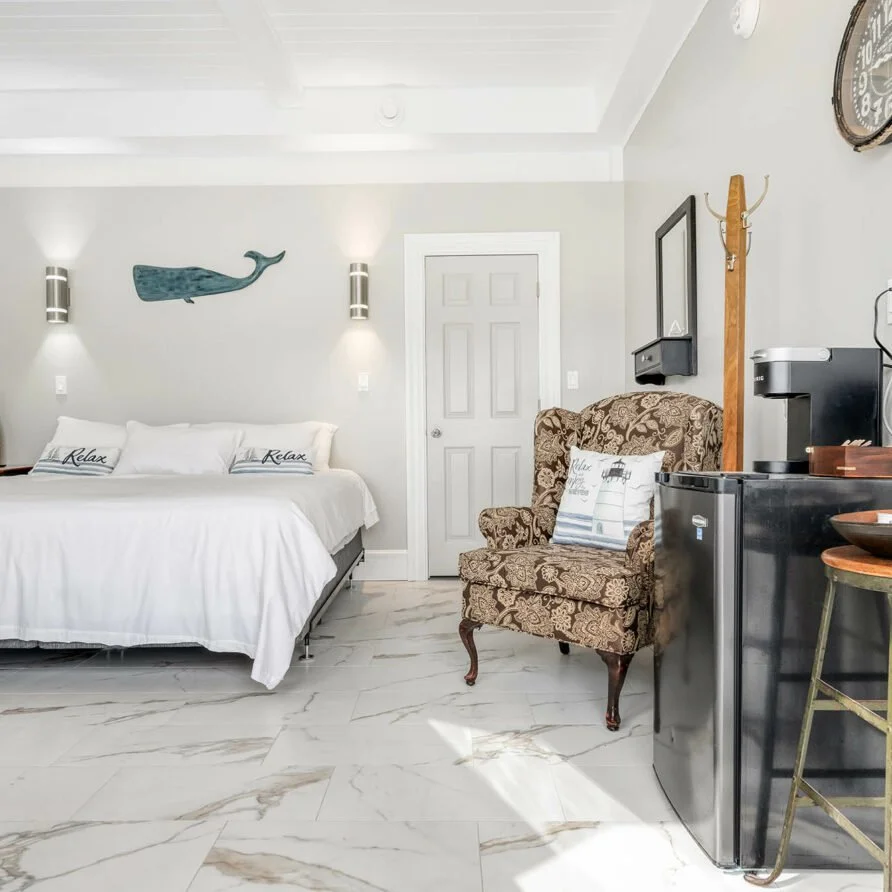 Interior view of a modern hotel room featuring a bed with white linens and pillows saying 'Relax', a patterned armchair with a pillow, a mini fridge with a coffee maker on top, and nautical decor including a whale and a lighthouse.