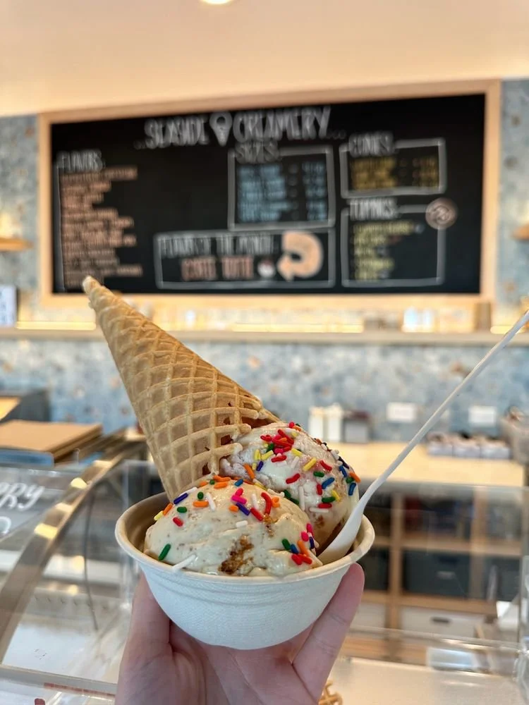 A cup of ice cream with rainbow sprinkles and a waffle cone on top, held in a person's hand at an ice cream shop with a chalkboard menu in the background.