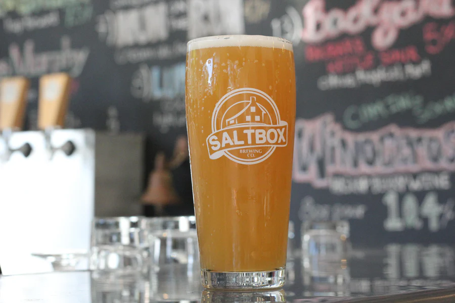 A pint glass of hazy, golden craft beer with a white foam head, featuring the Saltbox Brewing logo, on a bar counter with a chalkboard menu in the background.