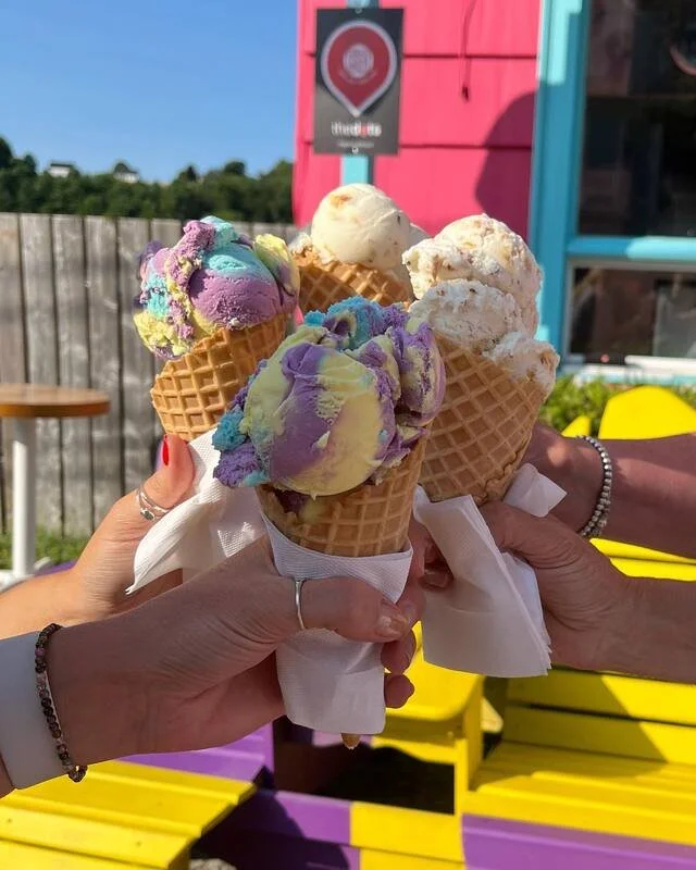 Three people holding ice cream cones with colorful and vanilla flavored ice cream outside on a sunny day.
