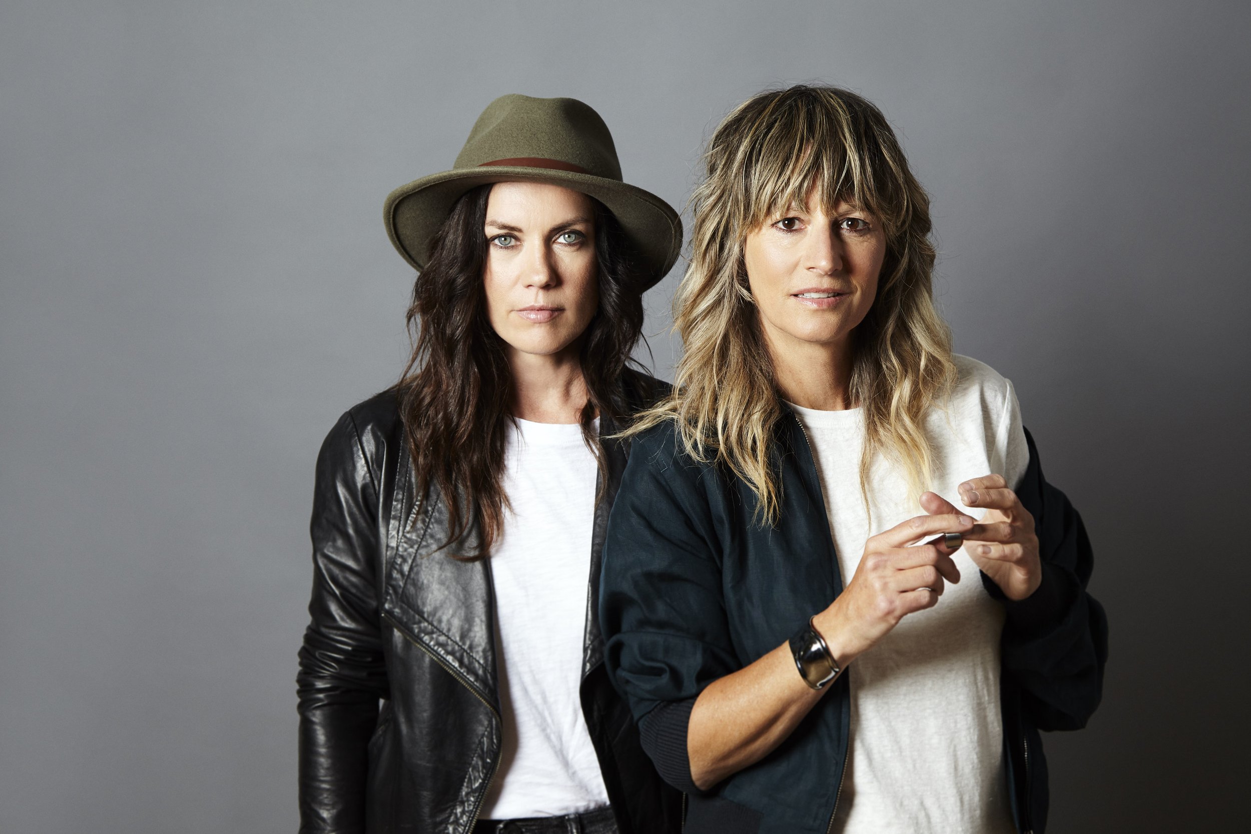 Two women with wavy hair, dressed in casual jackets and t-shirts, standing against a plain grey background.