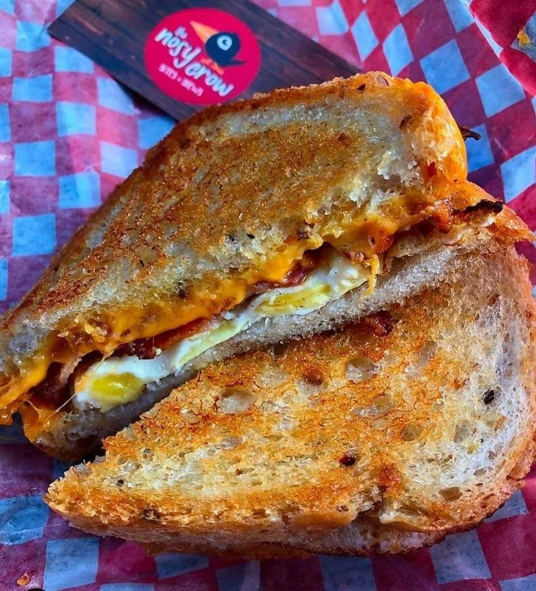 Close-up of a breakfast sandwich with fried egg, cheese, and bacon in toasted bread on red checkered paper.