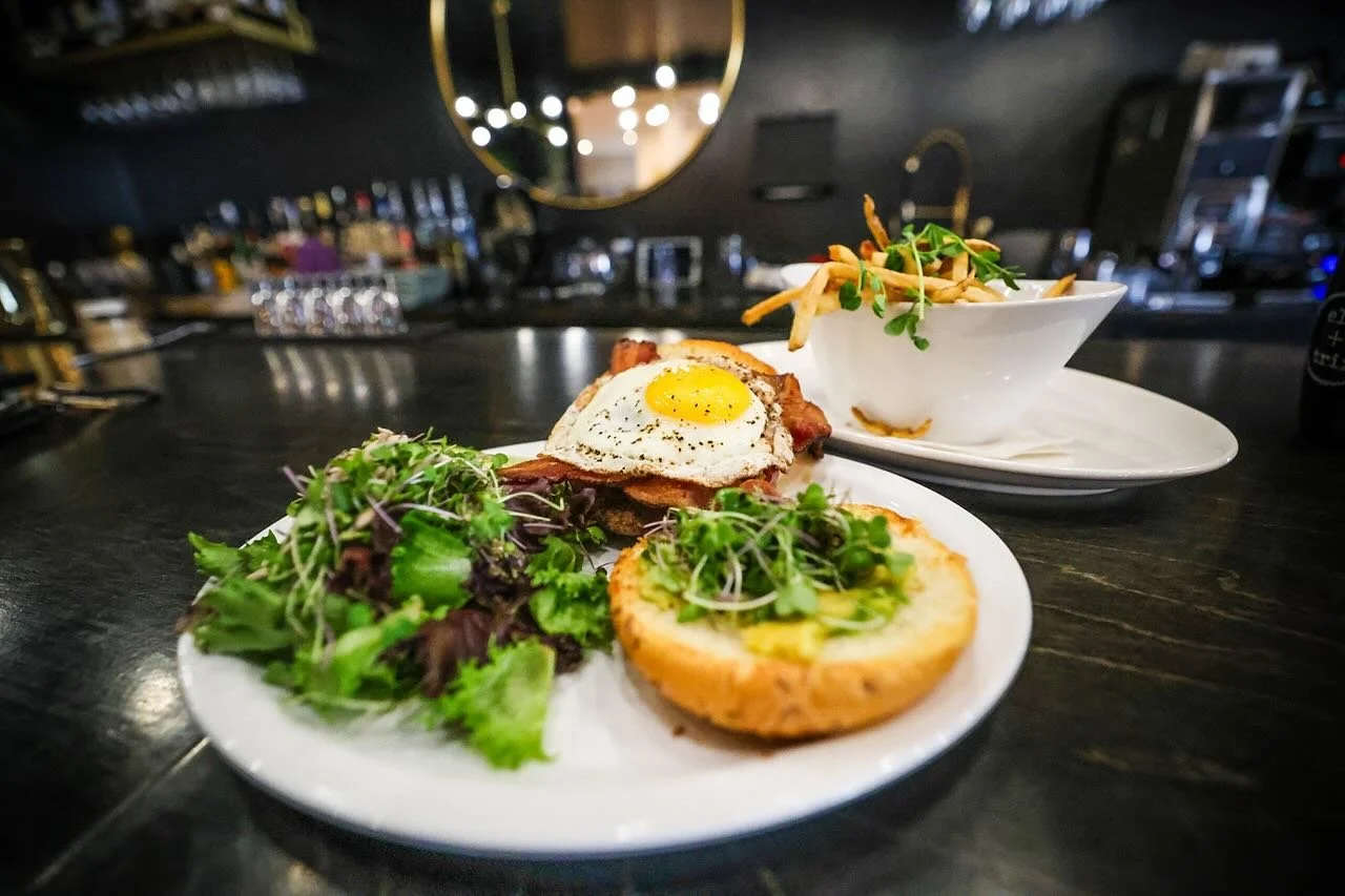A plate with a salad, a slice of toast topped with greens, and a fried egg with bacon. In the background, there is a bowl of fries garnished with microgreens, on a white plate, on a dark bar counter.