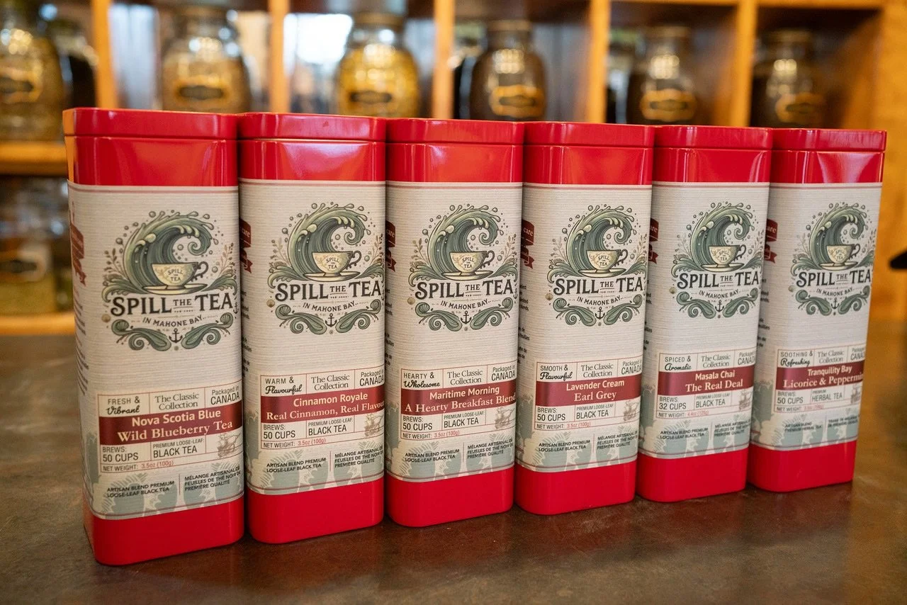 Six tins of Spill the Tea flavored black tea on a wooden surface with a blurred background of jars on a shelf.
