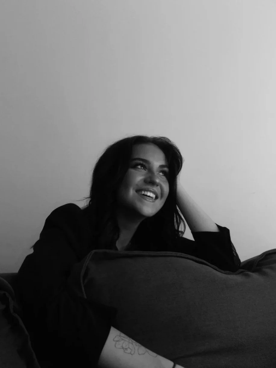 A woman with dark, wavy hair, smiling and looking up, resting her head on her hand, sitting behind a large cushion or pillow, in black and white.