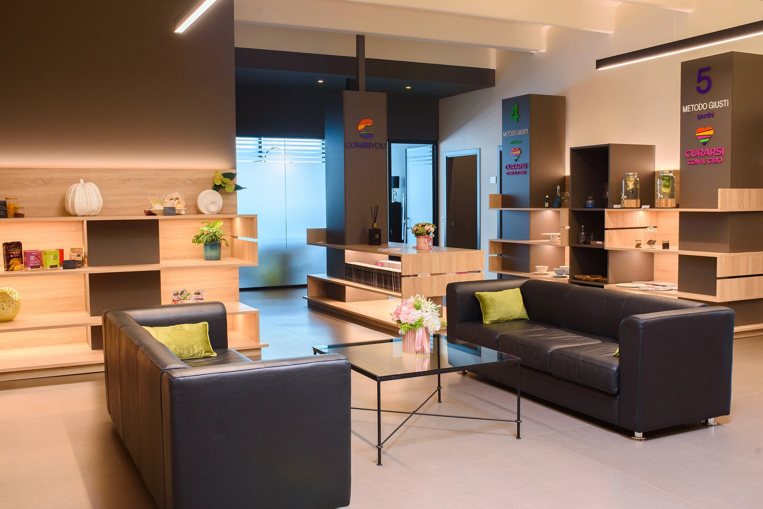 Interior moderno con due divanetti neri, un tavolino con vaso di fiori rosa, scaffali di legno con decorazioni e oggetti, pareti con numeri e messaggi colorati, ambiente accogliente e ben illuminato.