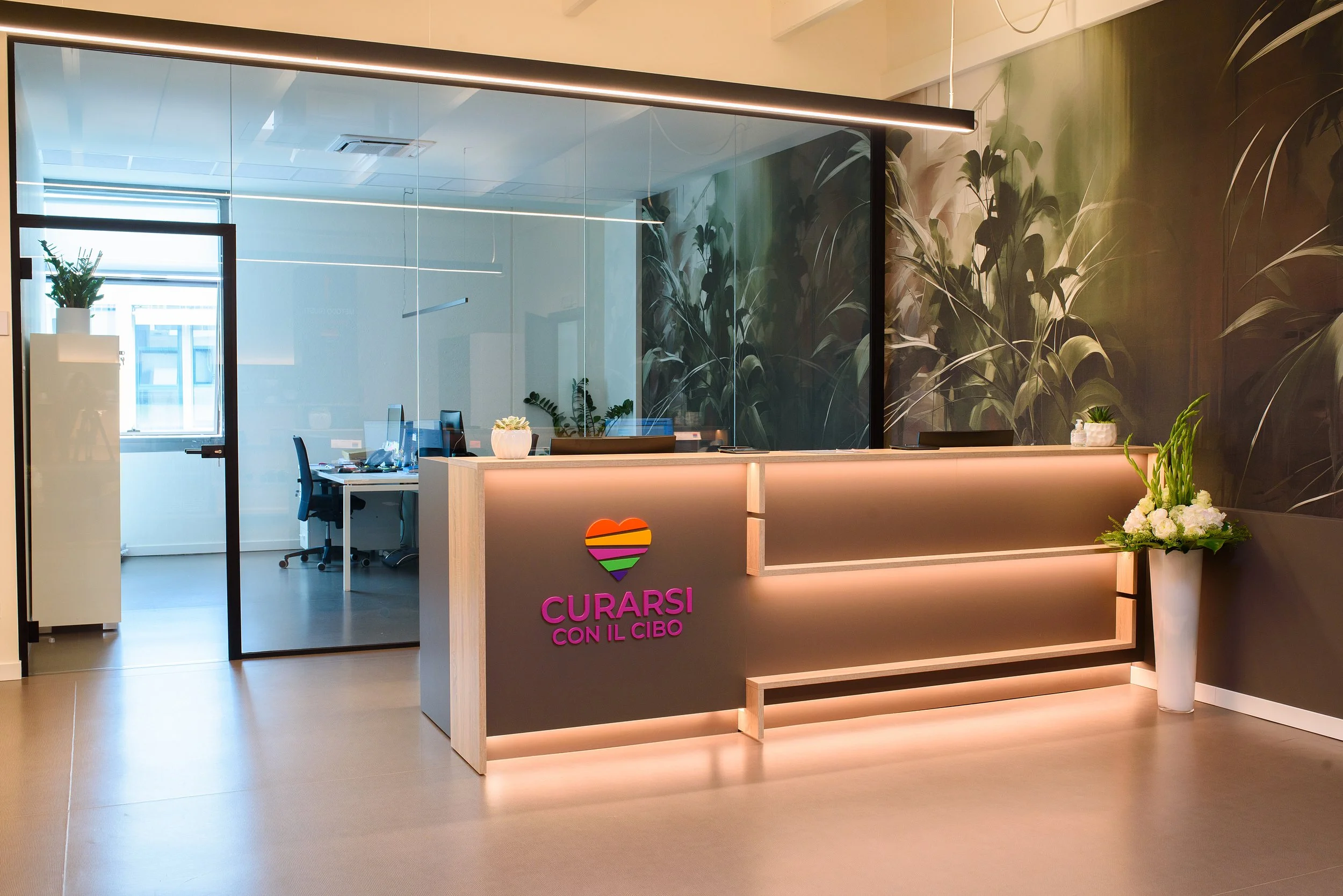 Reception moderna con logo con cuore arcobaleno e scritte 'Curarsi con il cibo', decorata con piante e fiori, con uffici visibili attraverso una parete di vetro.