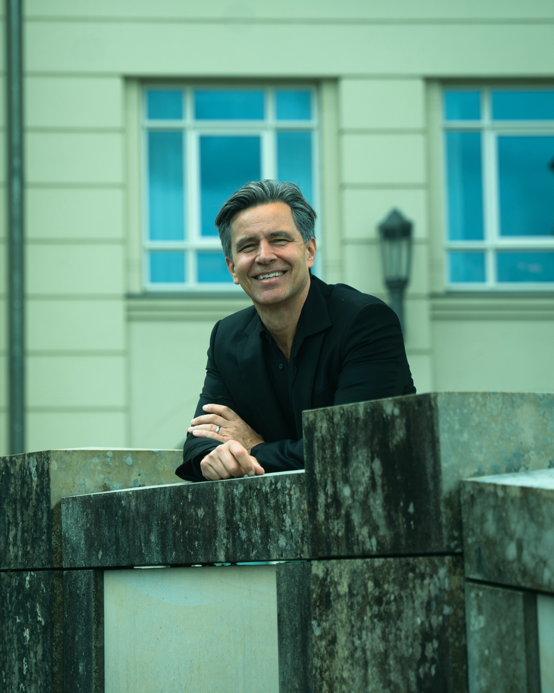 A smiling man in a black suit leaning on a stone wall with a background of a building with large windows and a lamp post.