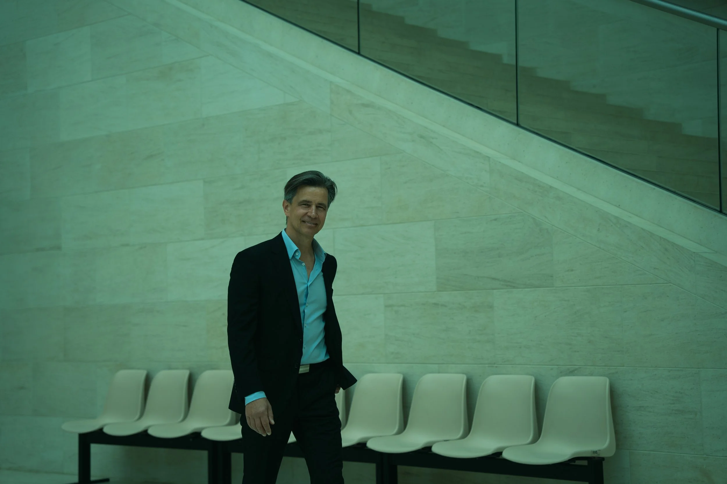 A man in a dark suit and light blue shirt standing in a modern building lobby with beige chairs and a green-tinted wall.