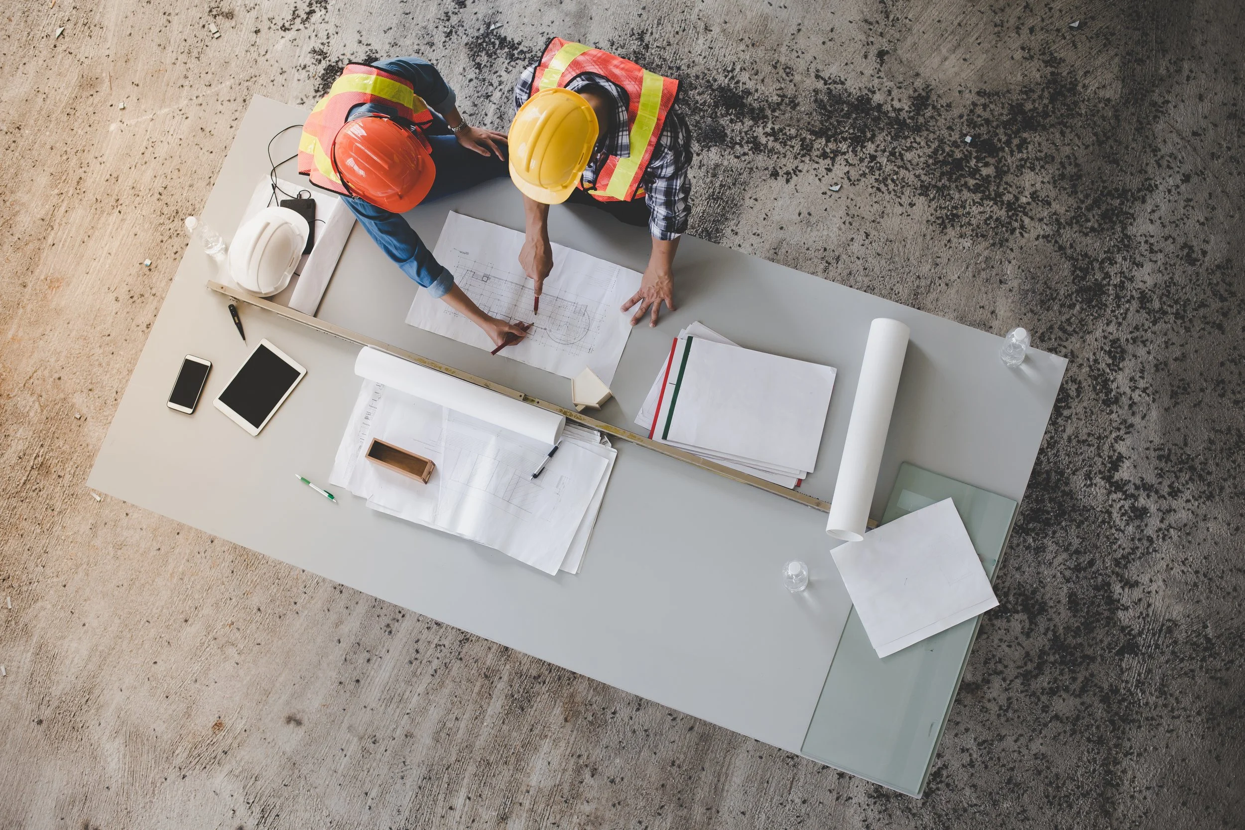 Two construction workers wearing helmets and safety vests are discussing blueprints on a table at a construction site.