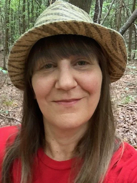 Woman with long brown hair wearing a striped straw hat and a red shirt, smiling outdoors in a wooded area.