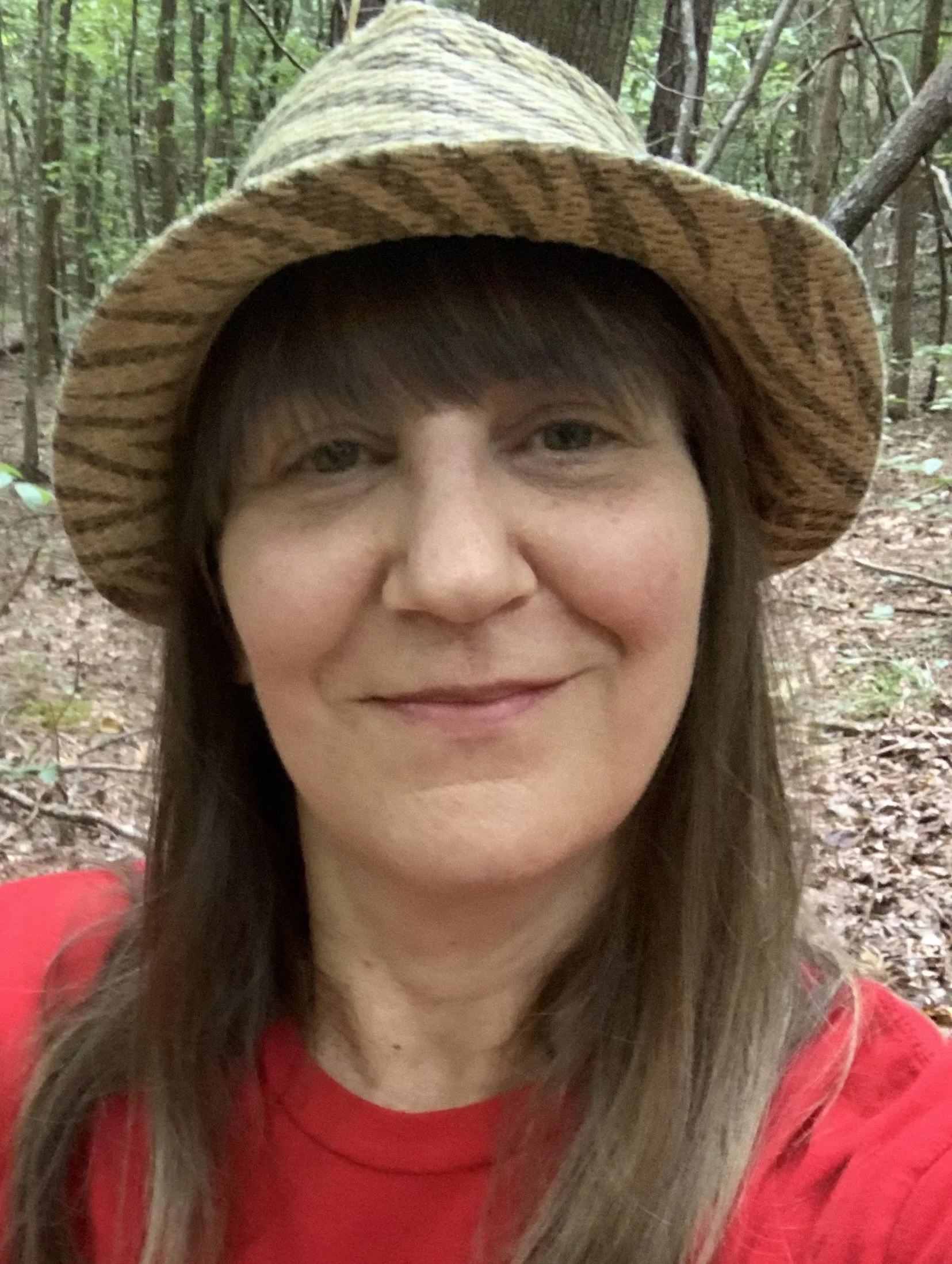 A woman wearing a straw hat and red shirt taking a selfie in a wooded forest.