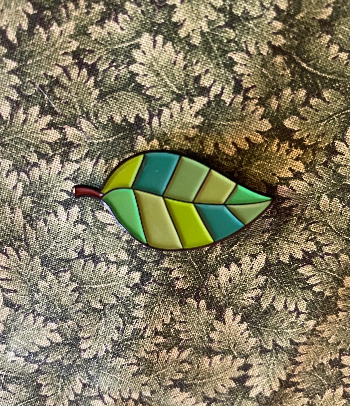 Colorful enamel leaf-shaped pin with green, yellow, and blue sections on a patterned fabric background.