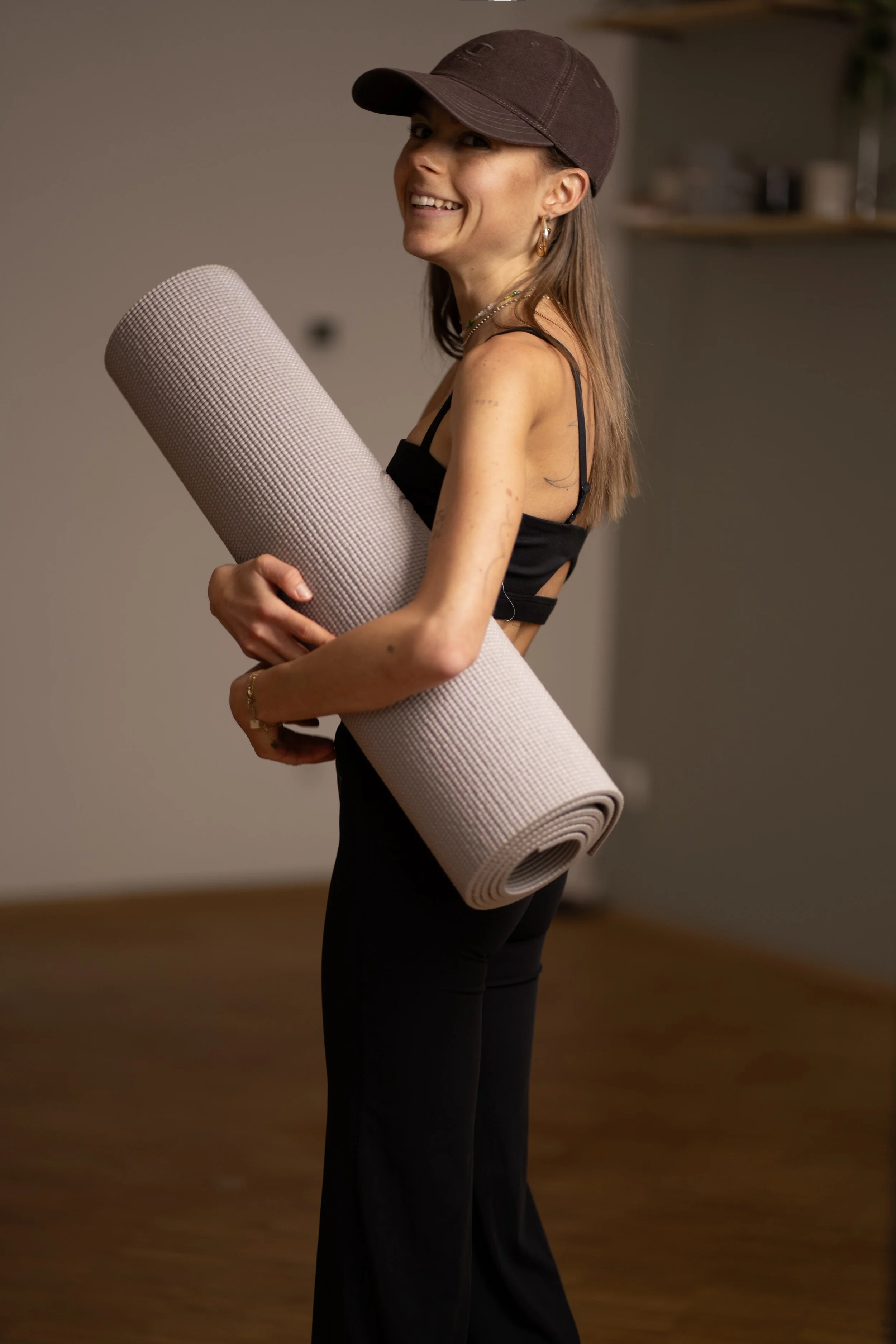 Eine lachende Frau mit Cappie und Schmuck, die einen Yogamatte in den Armen hält, steht in einem Raum mit Holzboden.