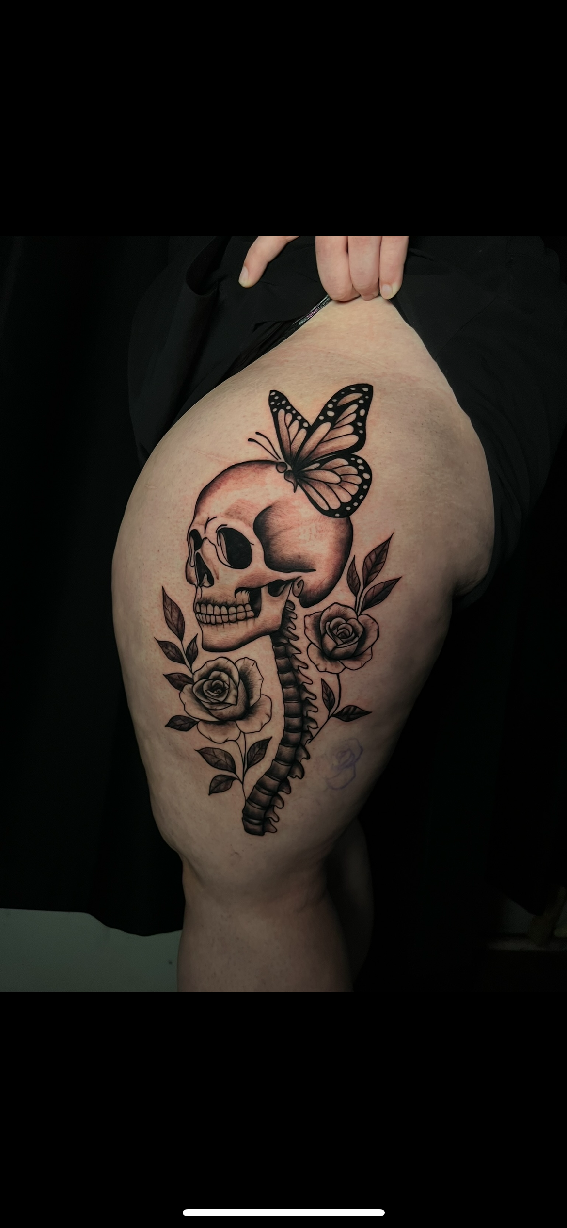 Tattoo of a human skull with a butterfly on top and roses with leaves around it on a person's thigh.