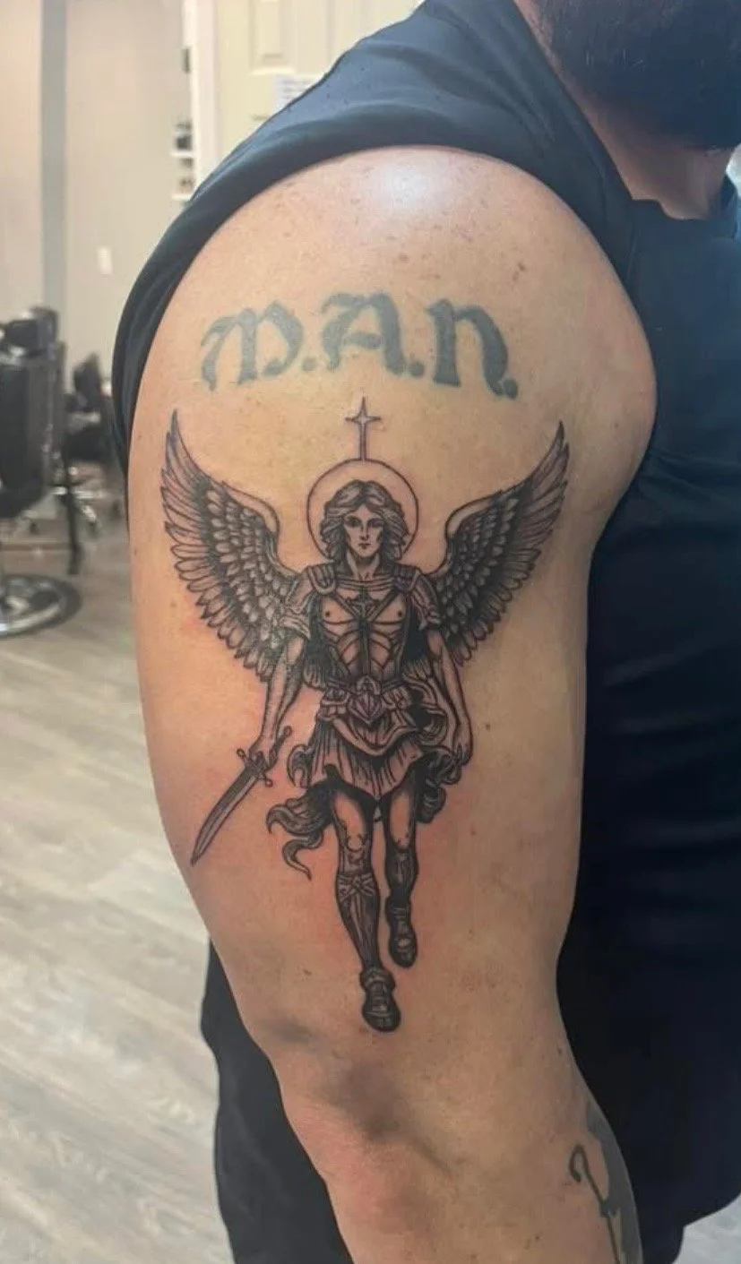 Tattoo of a winged female warrior with a sword, a halo, and armor on upper arm; the word 'M.A.R.' in stylized letters above her.