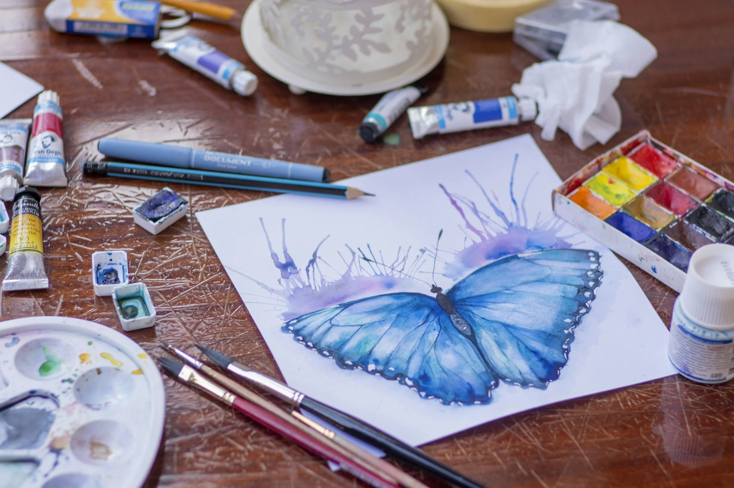 A watercolor painting of a blue butterfly on paper surrounded by paint tubes, brushes, watercolors, and art supplies on a wooden table.