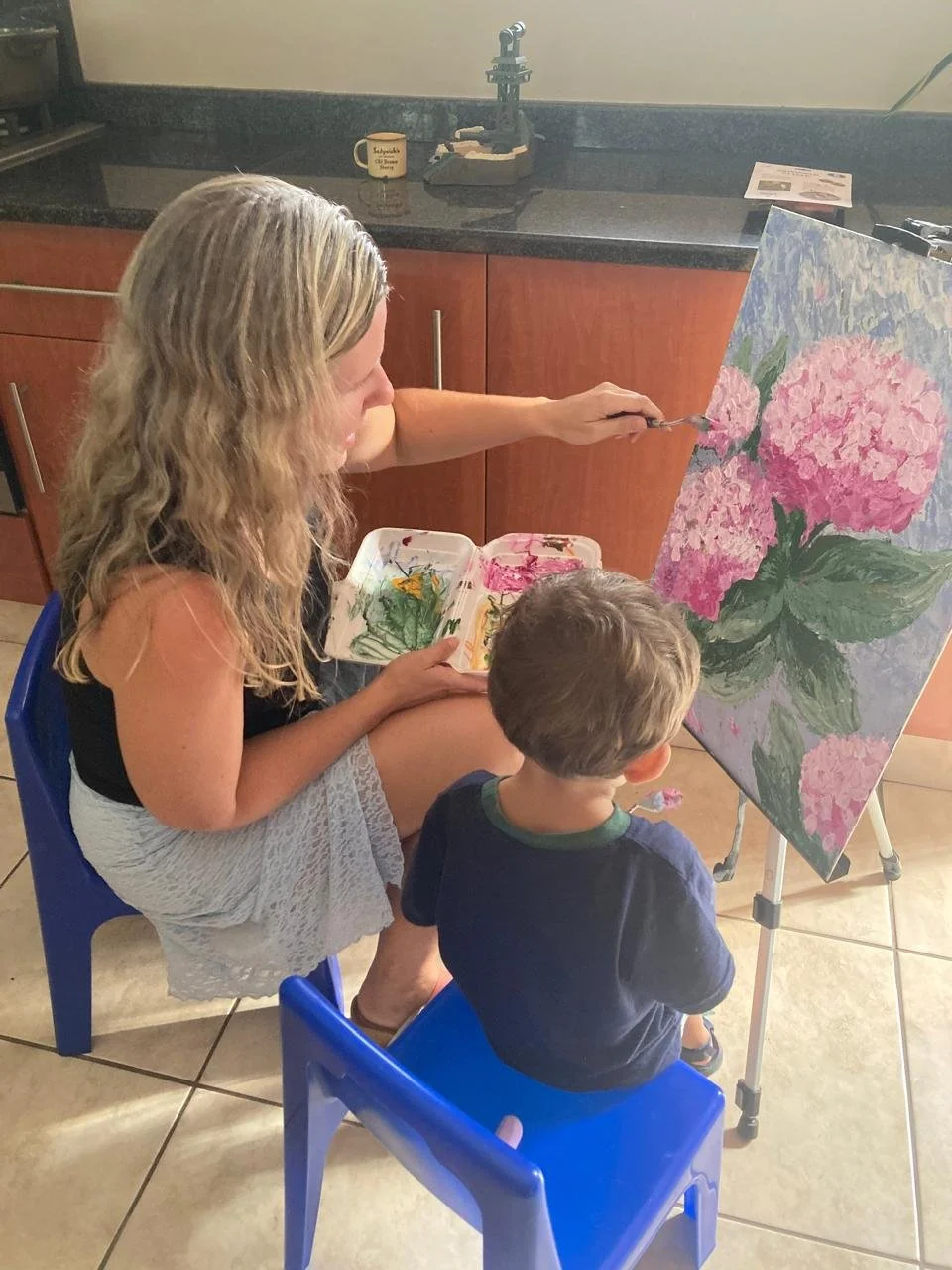 Tracy Marais is painting a large canvas of pink hydrangea flowers with her 2 year old son. Tracy is holding a palette and applying pink paint to the flowers. This artwork is acrylic and create with palette knives.