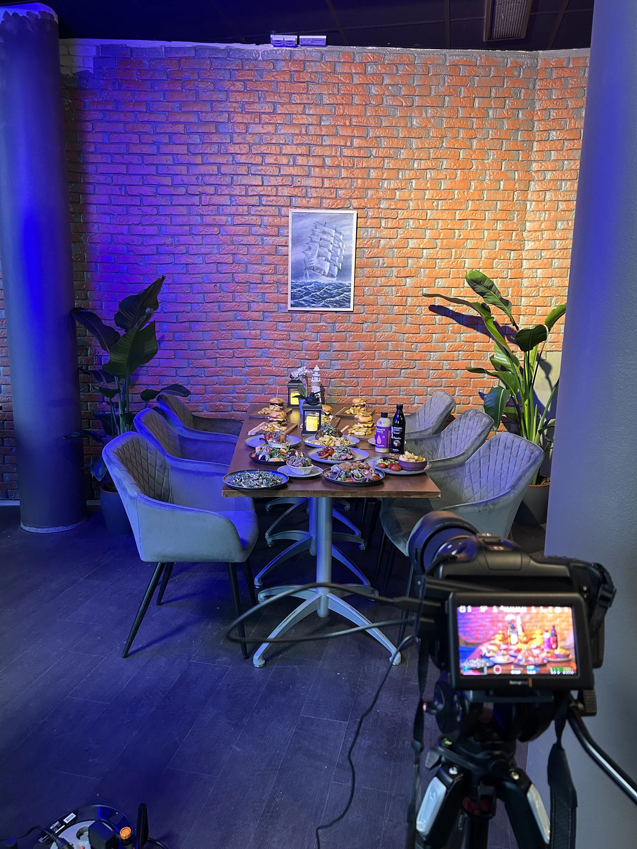 A dining table with plates of food in a restaurant with a brick wall, plants, and wall art. A camera's viewfinder is in the foreground.