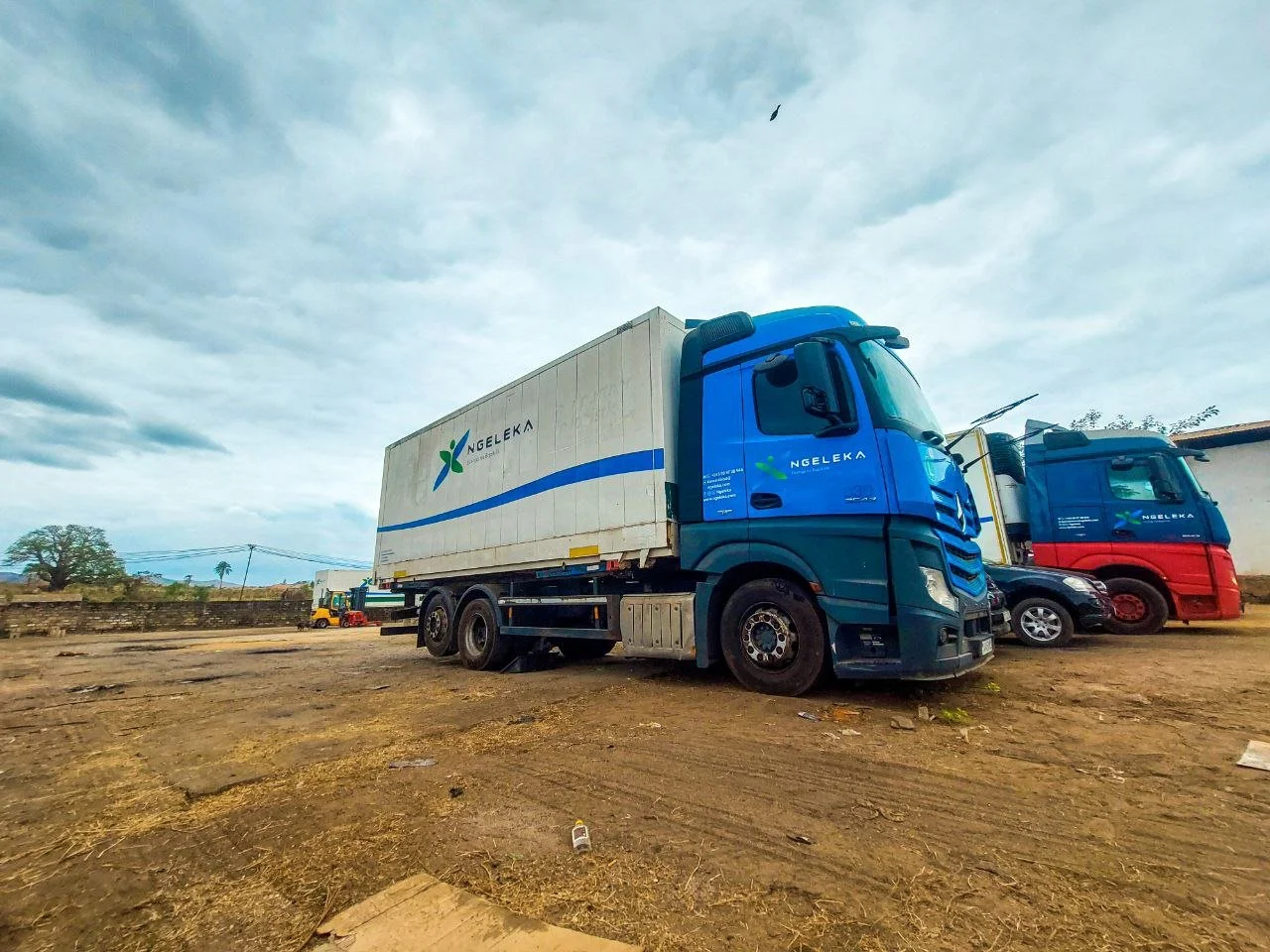 Camion de transport avec logo Ngeleka, stationné sur un terrain poussiéreux sous un ciel nuageux, avec un autre camion en arrière-plan et un arbre distinctif à gauche.