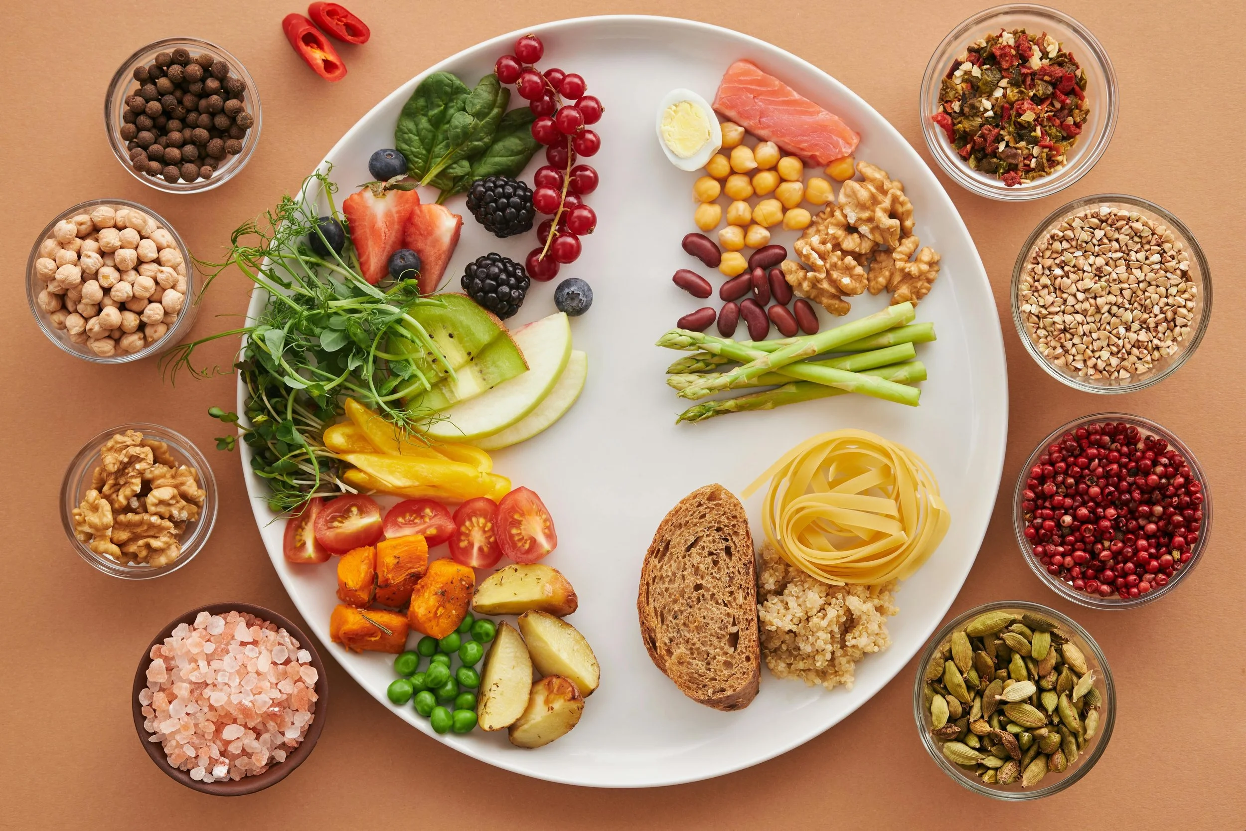 plate full of nutritious vegetables, fruits, fibers, and starches. the plate is full of colorful food representing a healthy diet with other small bowls of nuts and seeds surrounding it.