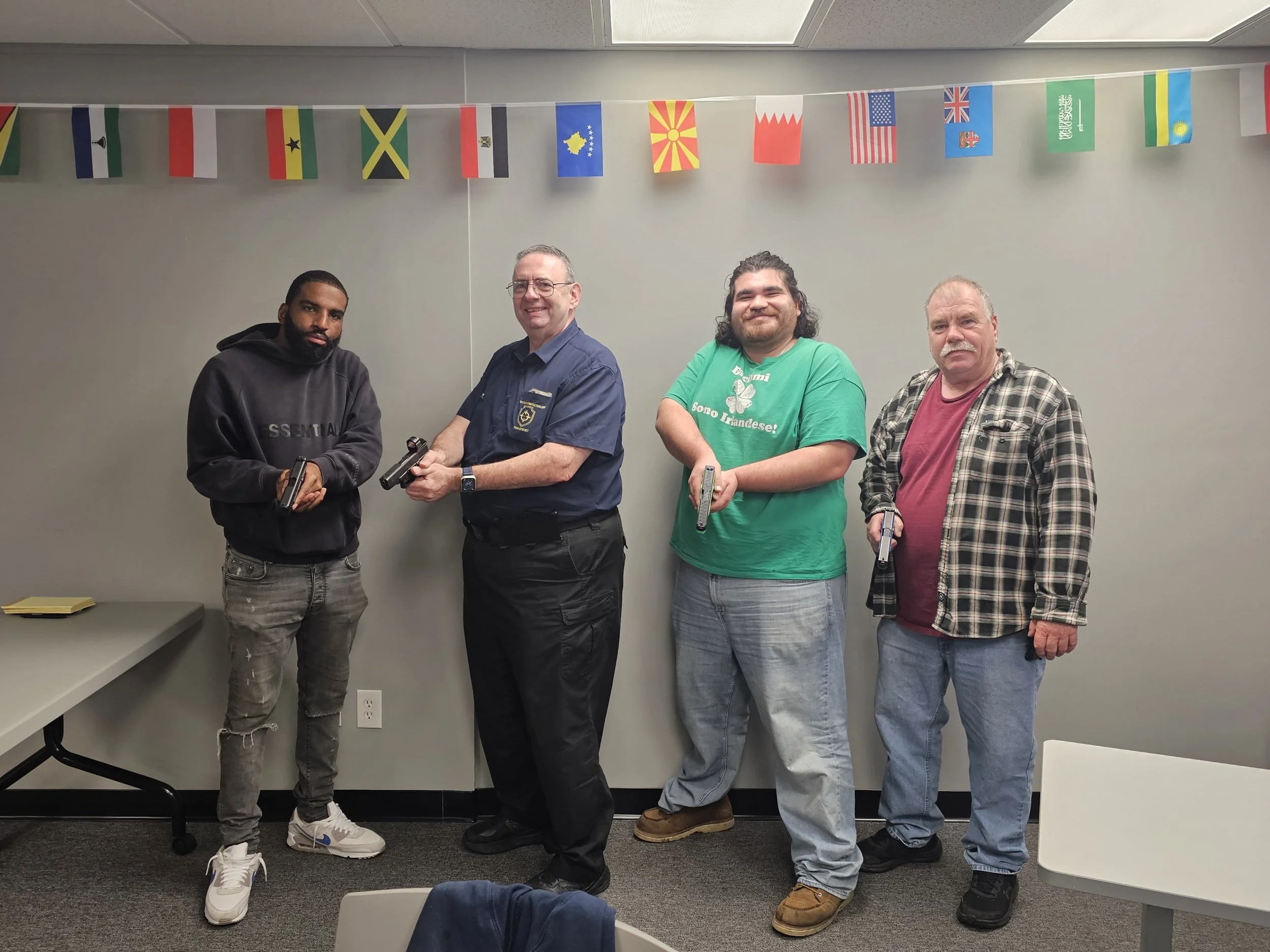 Four men standing indoors, holding guns, with a string of international flags hanging overhead.