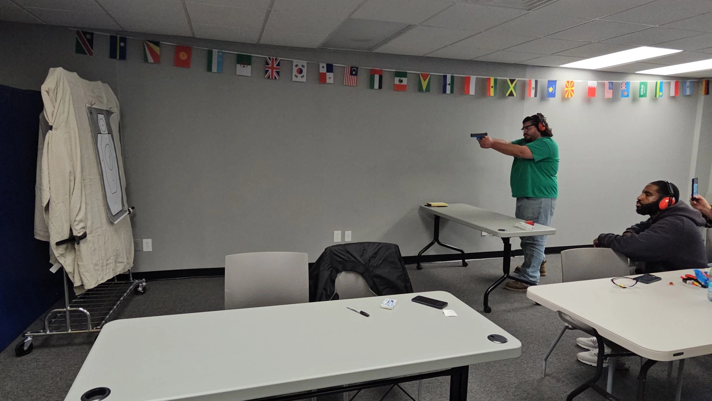 A man wearing glasses and a green shirt is aiming a gun at a shooting target in an indoor range. Another man with headphones is sitting at a table nearby, using a cellphone. The room has a gray wall decorated with small international flags hanging fr