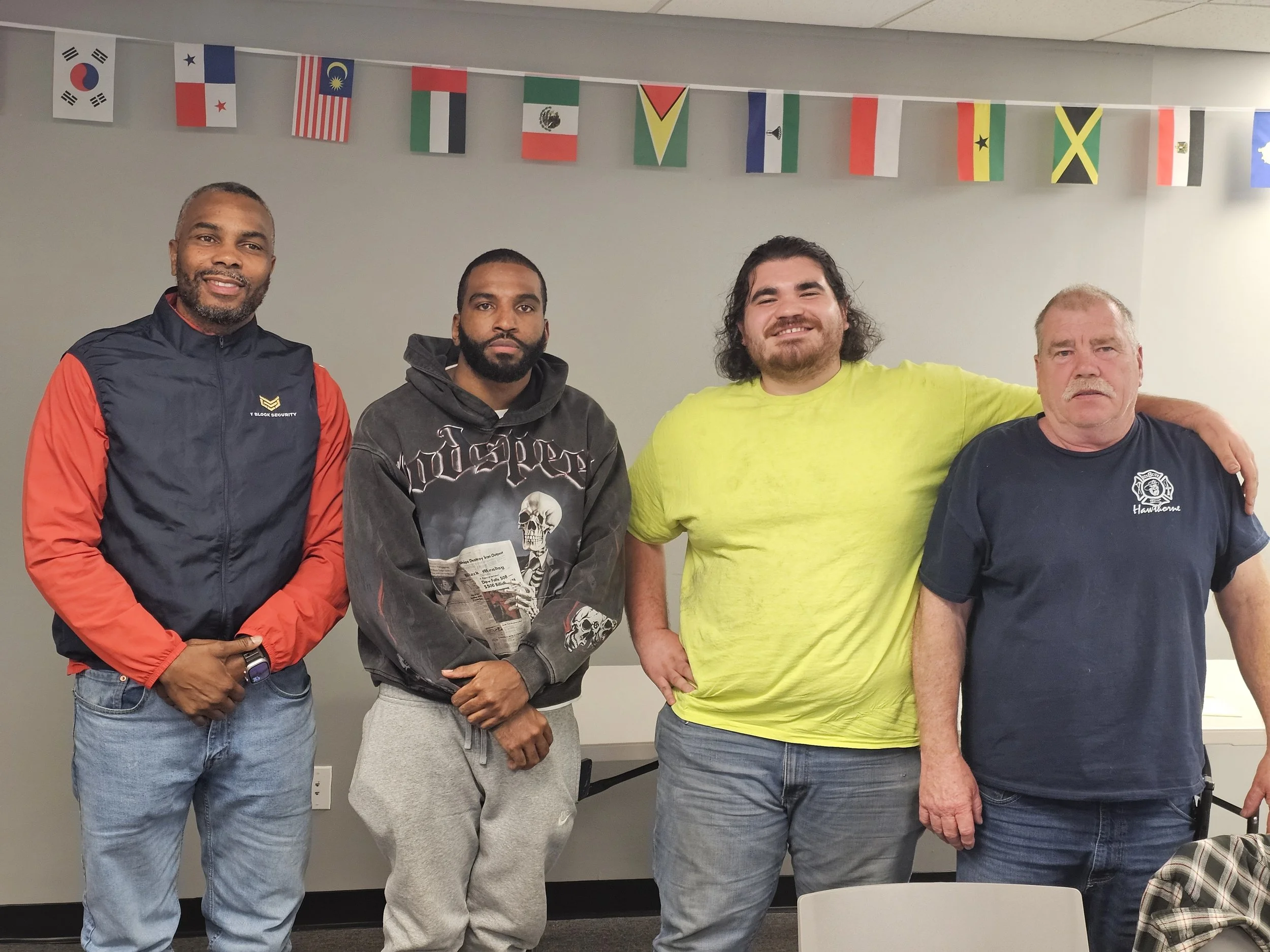 Four men standing indoors, with international flags hanging above them. The man on the far left is wearing a navy and red jacket, the second man is in a black hoodie with graphic art, the third man is in a bright yellow T-shirt, and the fourth man is