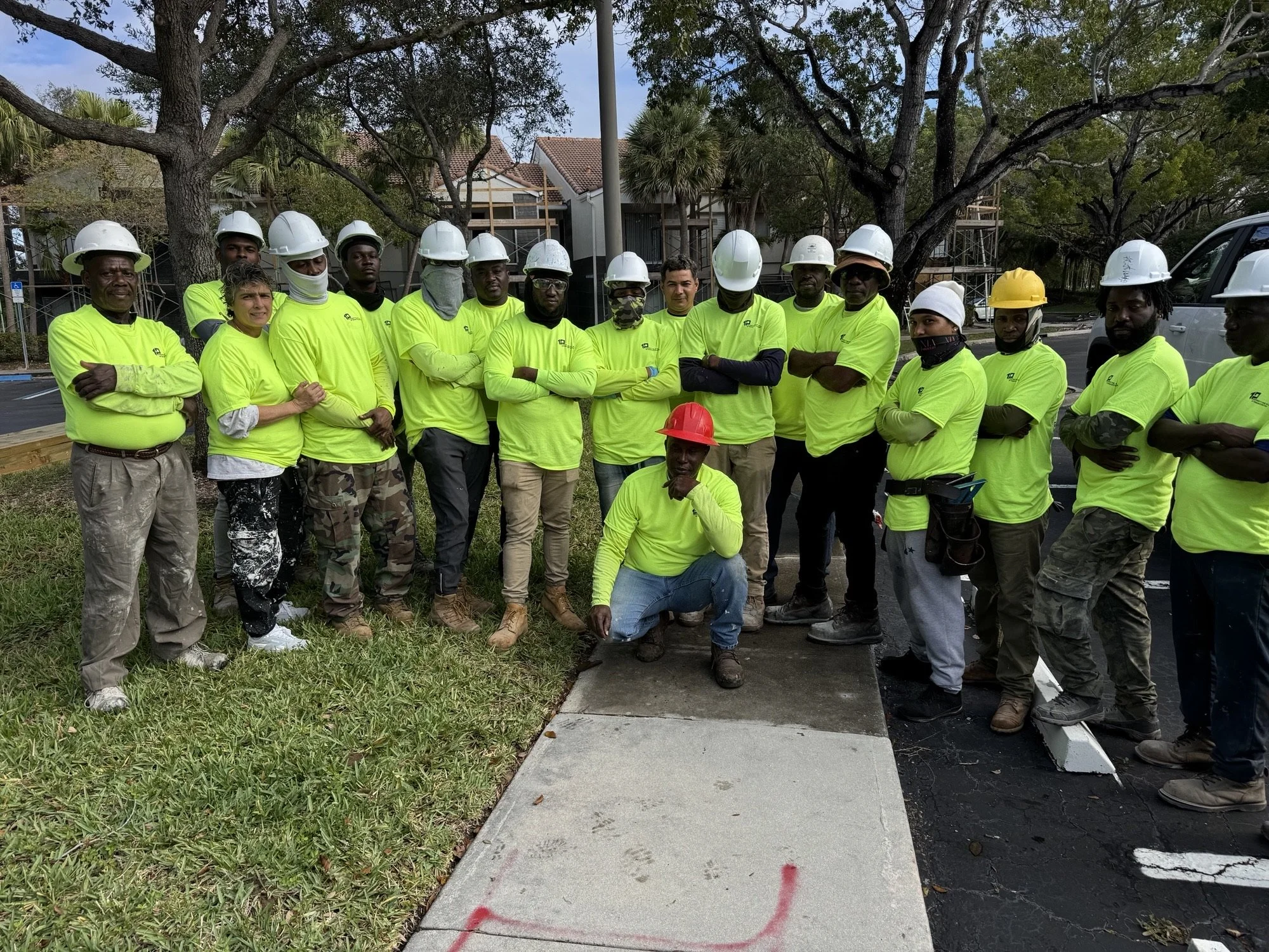 Construction workers wearing neon yellow shirts and various colored hard hats standing outdoors on a cloudy day, some with arms crossed, in front of trees, a building under construction, and a parked vehicle.