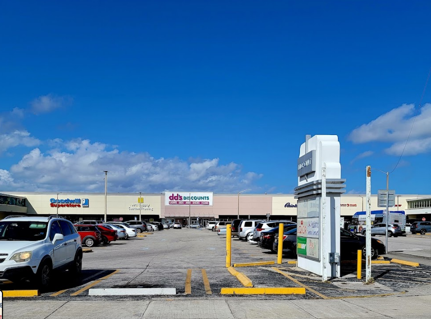 Parking lot in front of retail stores with a bright blue sky and scattered clouds overhead.