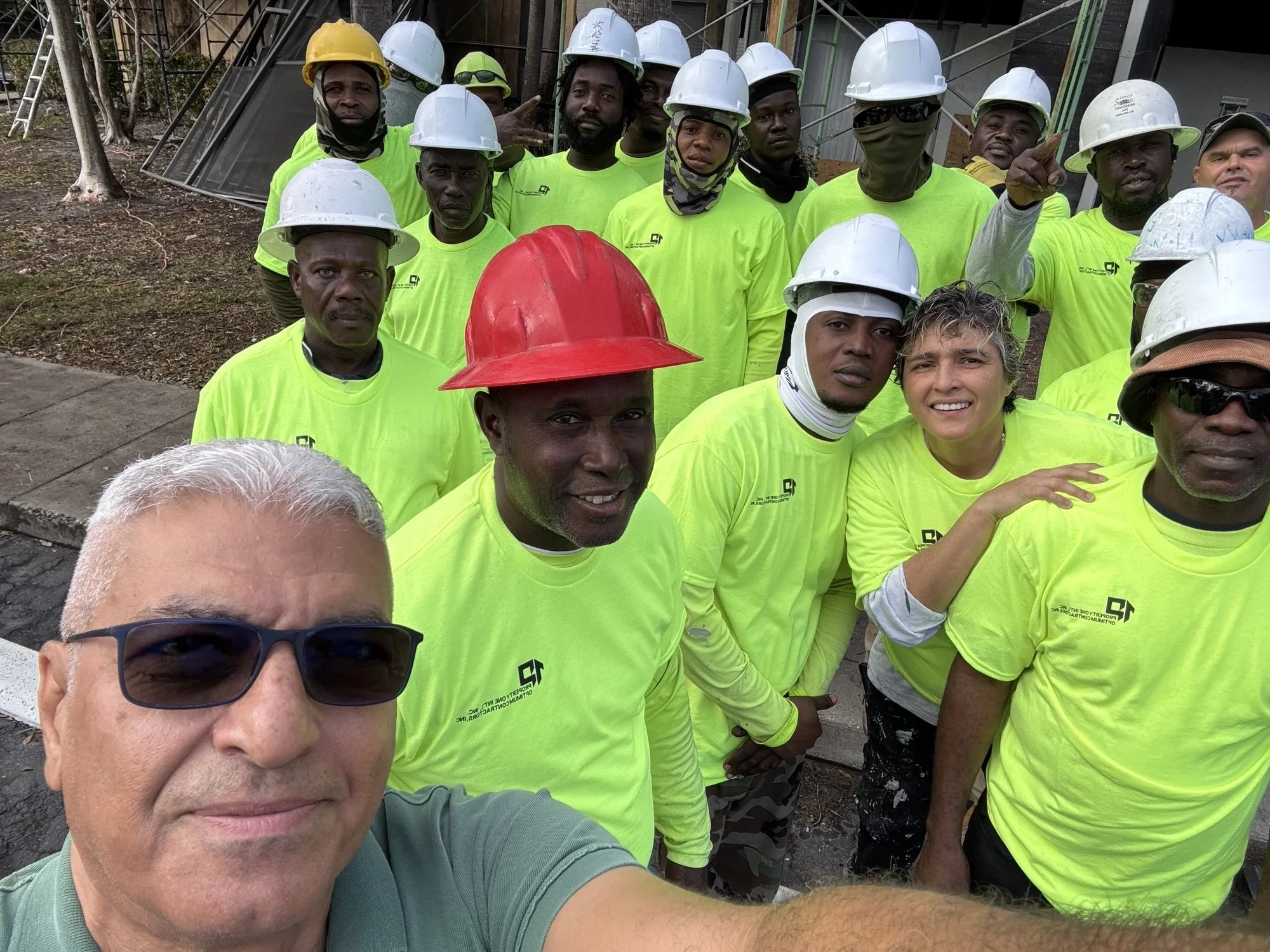 Group of construction workers wearing bright yellow shirts and hard hats, posing for a selfie on a construction site.