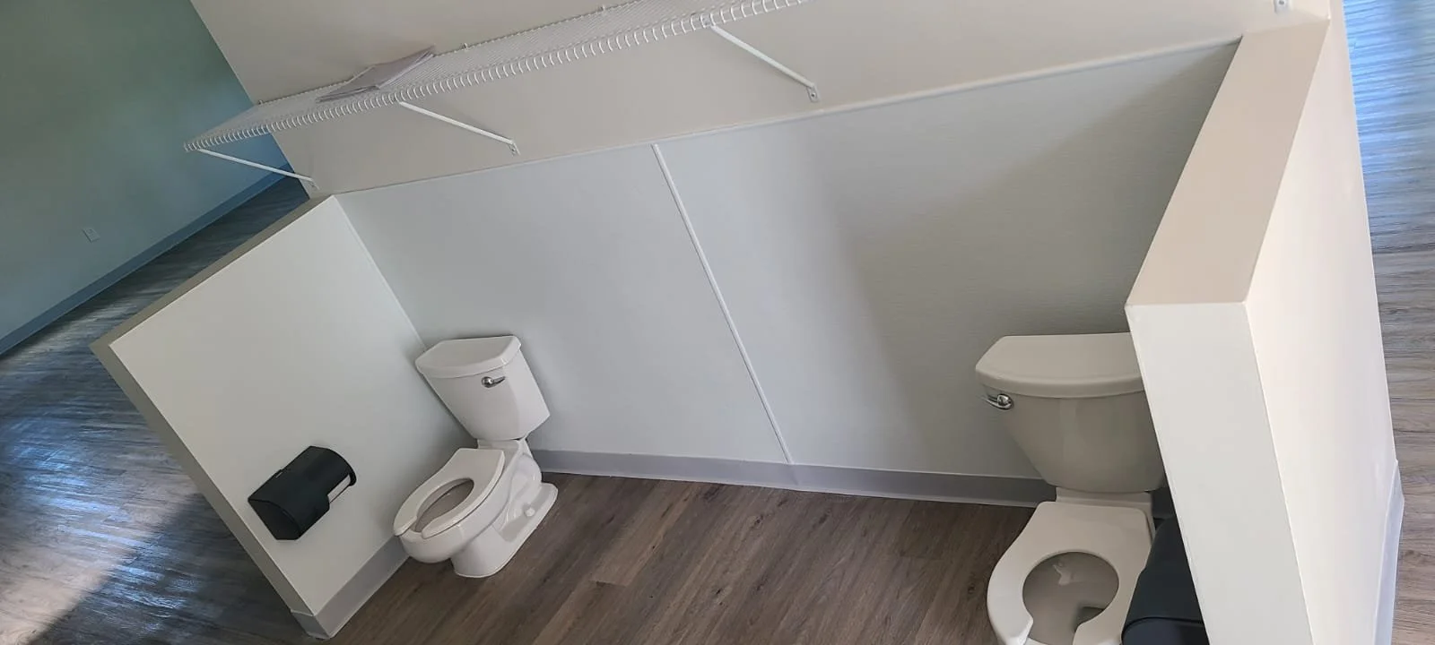 Small bathroom with two toilets separated by a half wall, one with a black trash bin and a shower rod with a shower curtain