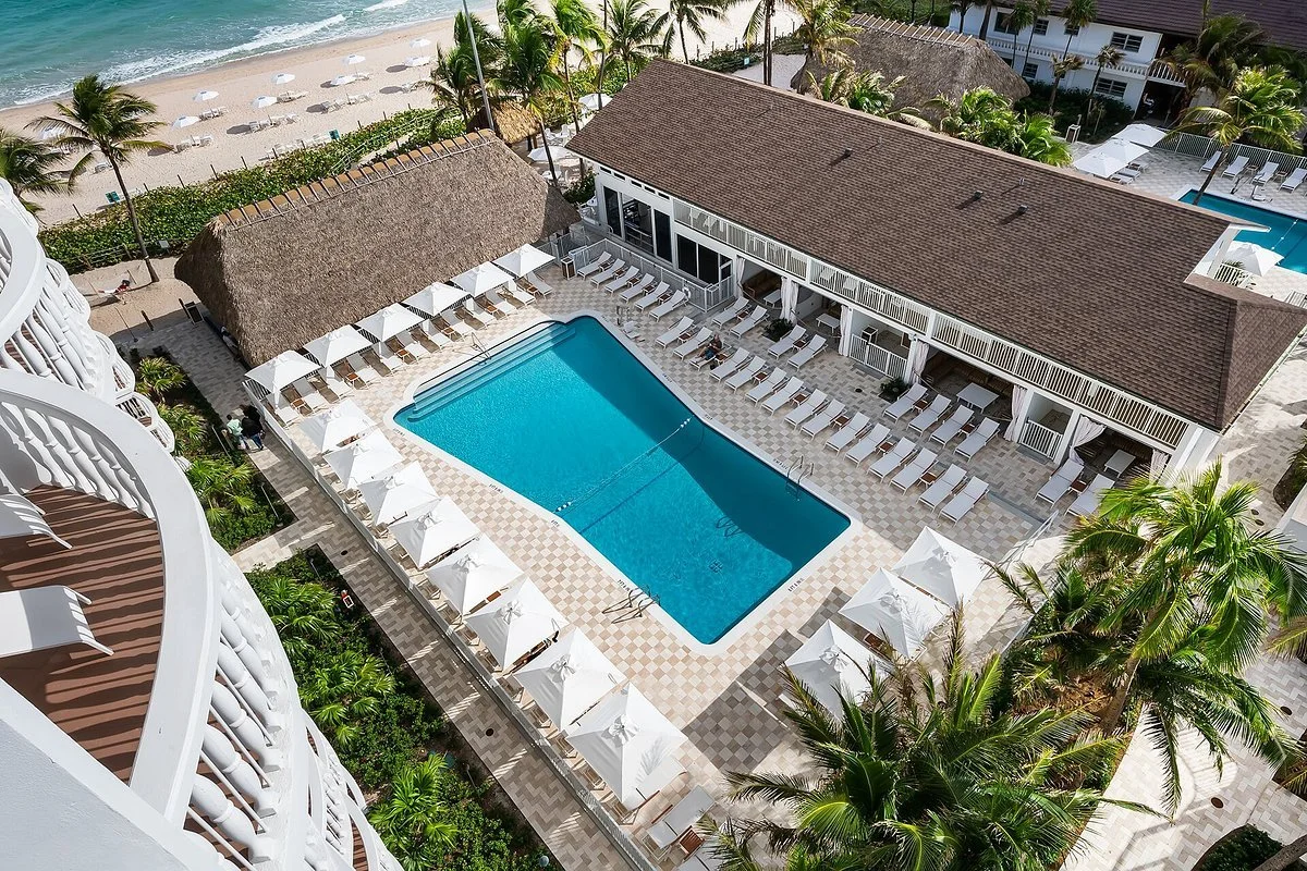 An aerial view of a pool area at a beachside resort with a swimming pool, white lounge chairs, white umbrellas, a thatched-roof hut, a building with balcony rooms, palm trees, sandy beach, and ocean in the background.
