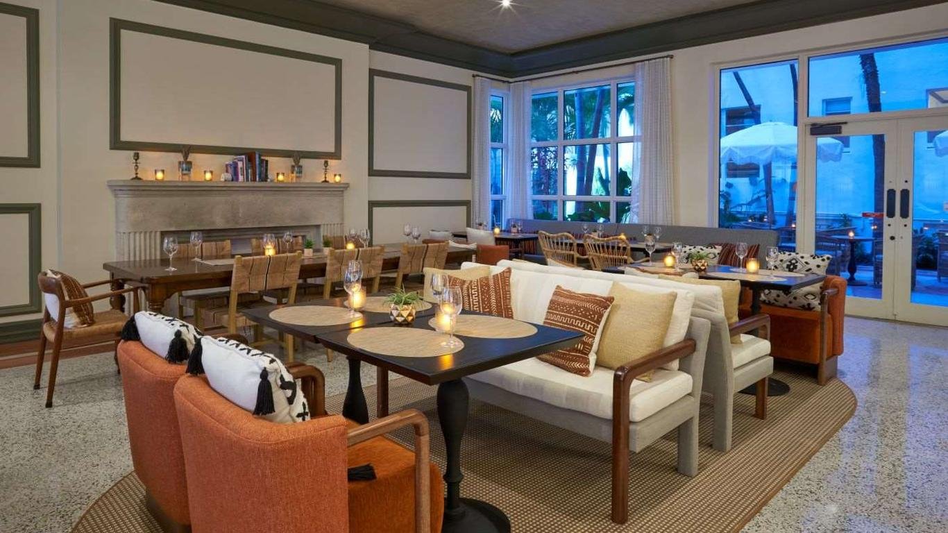 Elegant dining area with a large table, surrounded by mixed chairs and a sofa, illuminated by candlelight, with large windows showing an outdoor patio with palm trees and umbrellas.