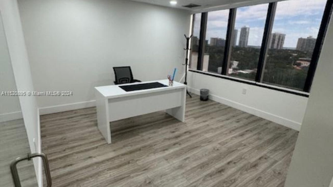 Empty modern office with white desk, black office chair, coat rack, large front window showing city and greenery, wood flooring, and white walls.