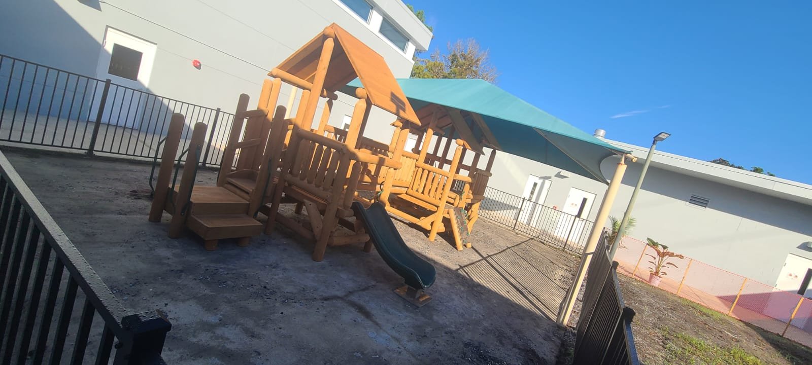 Children's playground with a wooden play structure, slide, and a shade canopy, enclosed by black fencing, next to a modern white building with small windows, under a clear blue sky.