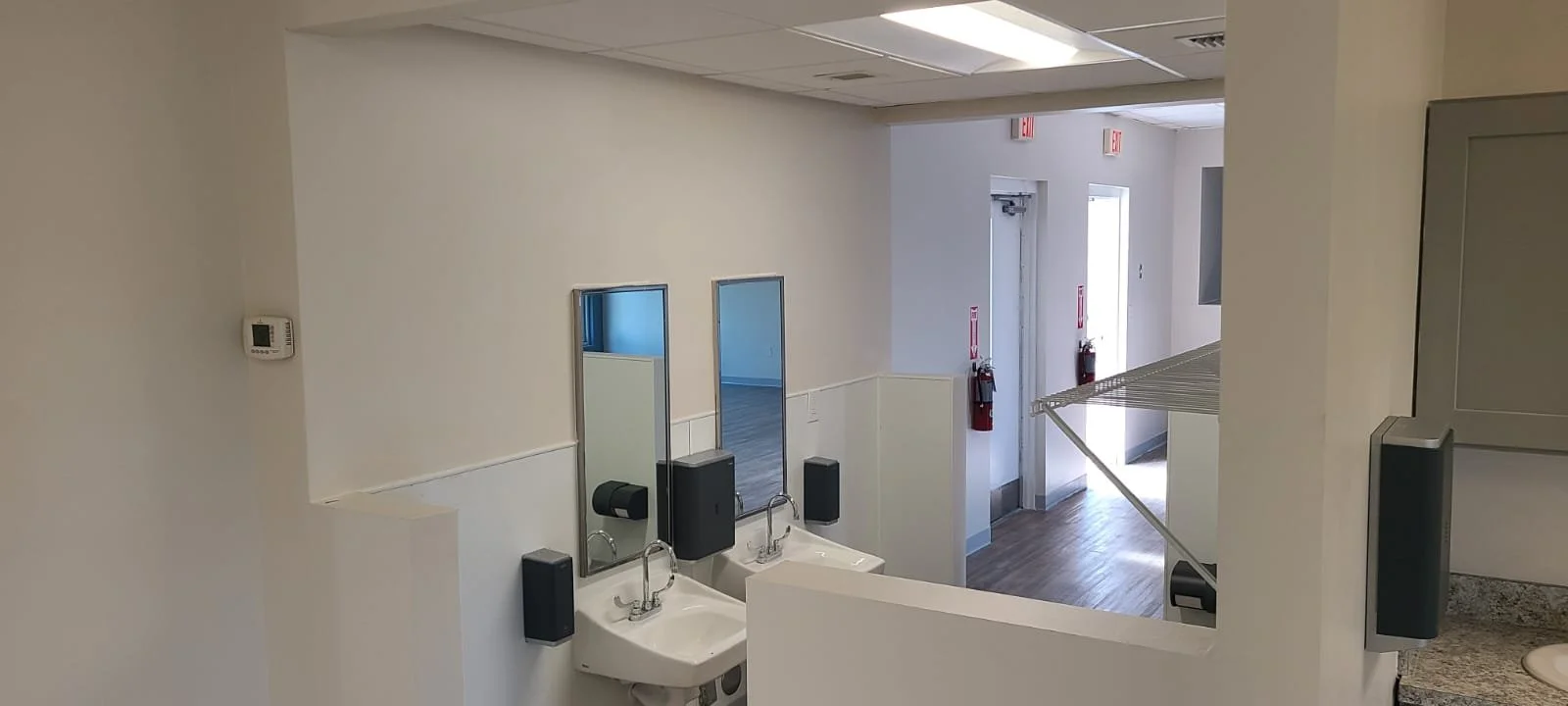Public restroom with two sinks, two mirrors, and soap dispensers, without users, showing part of a cleaning or storage area on the right.