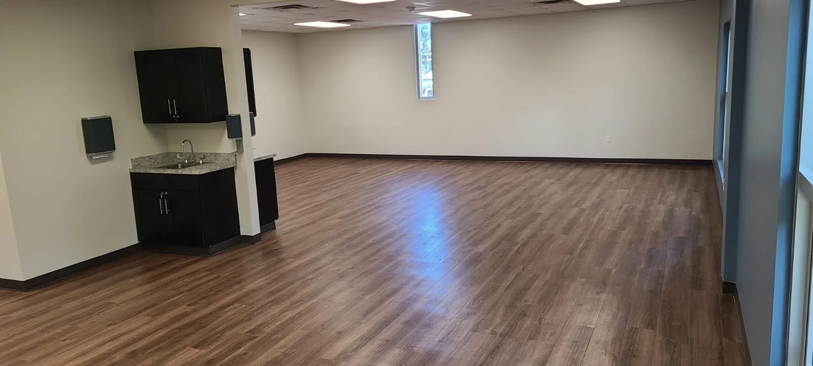 Empty room with hardwood floors, small kitchenette with sink, dark cabinets, and a narrow window.