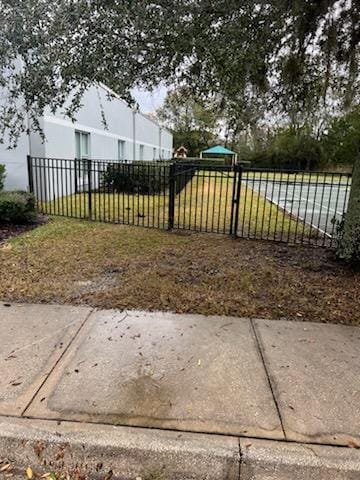 A sidewalk in the foreground with a small patch of dirt and grass, a black metal gate, and a grassy yard with residential buildings and trees in the background.