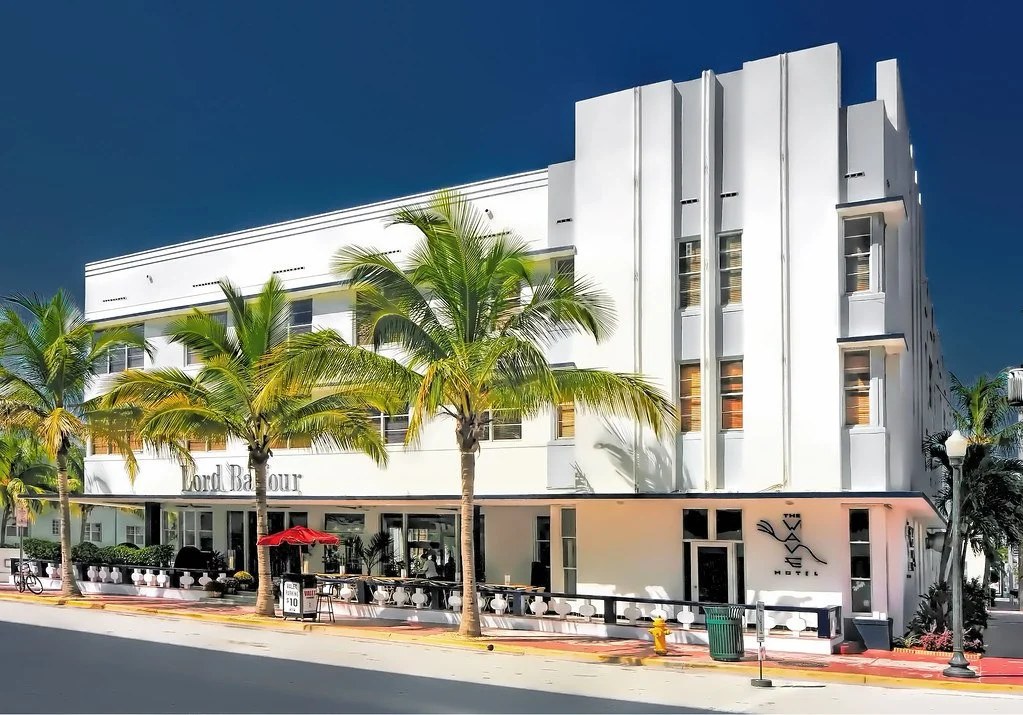 White Art Deco hotel building with palm trees and a red umbrella on a sunny day.