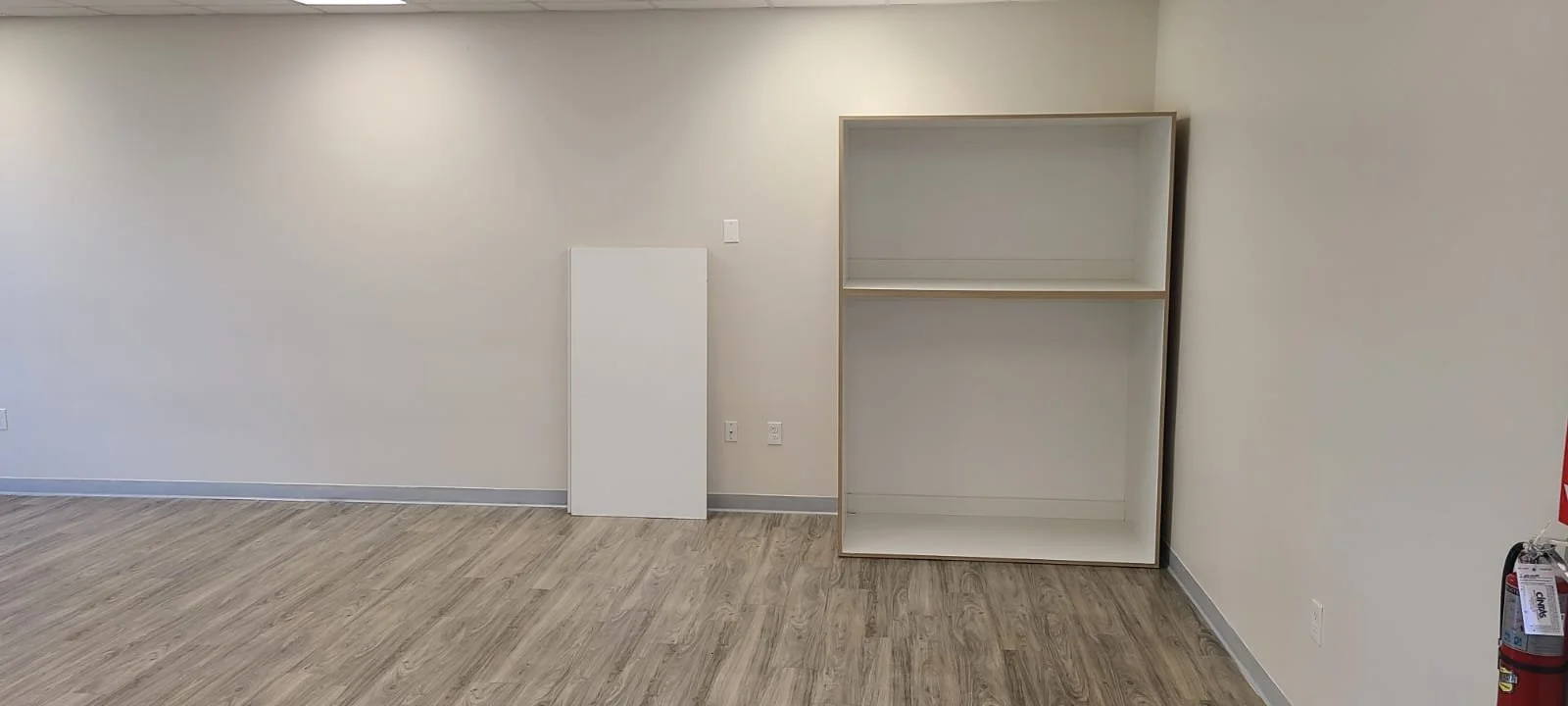 Empty room with beige walls, wood flooring, a white board leaning against the wall, a partially assembled white shelving unit, and a fire extinguisher in the corner.