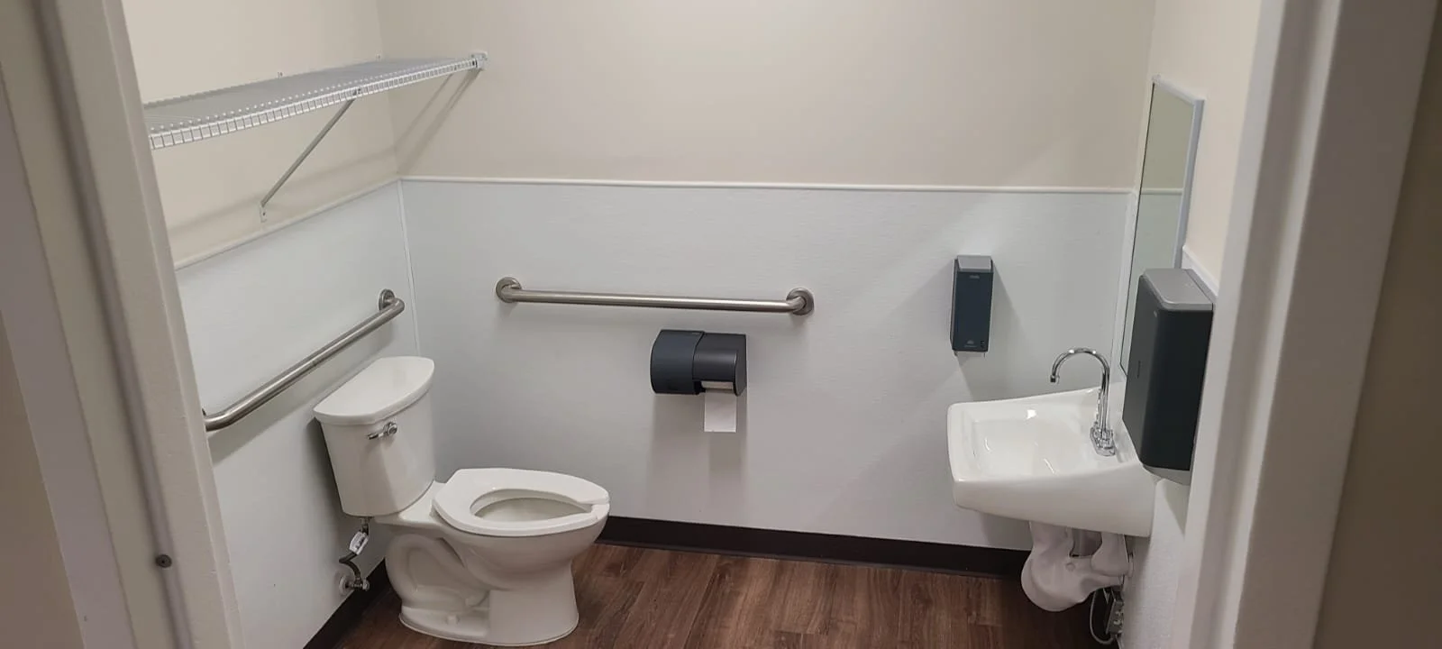 Accessible restroom with a toilet, small sink, mirror, soap dispenser, paper towel dispenser, and grab bars.