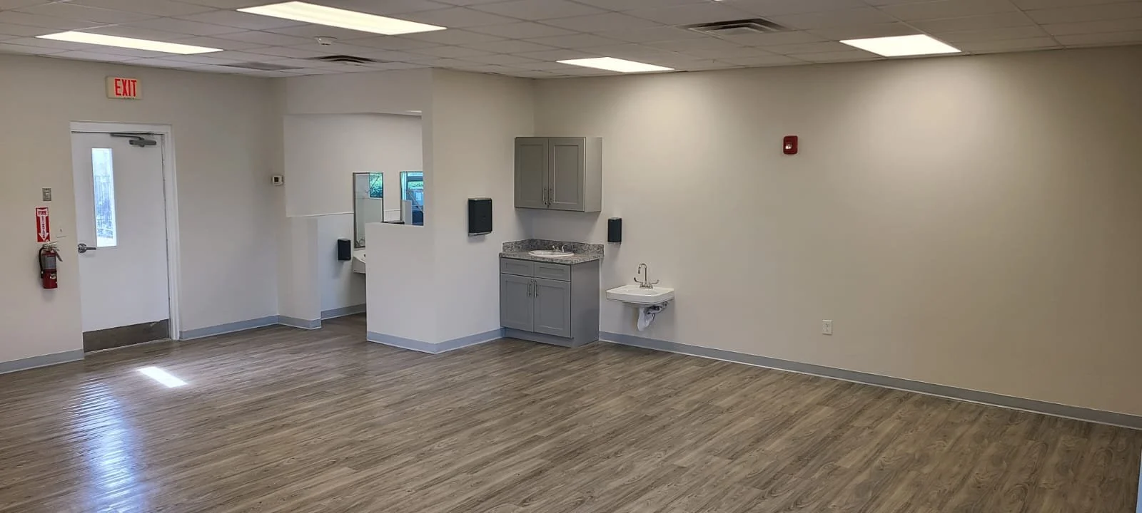 Empty room with wood-like flooring, a door on the left, a small sink with gray cabinets, a fire extinguisher on the wall, a fire alarm, and a windowed area in the background.
